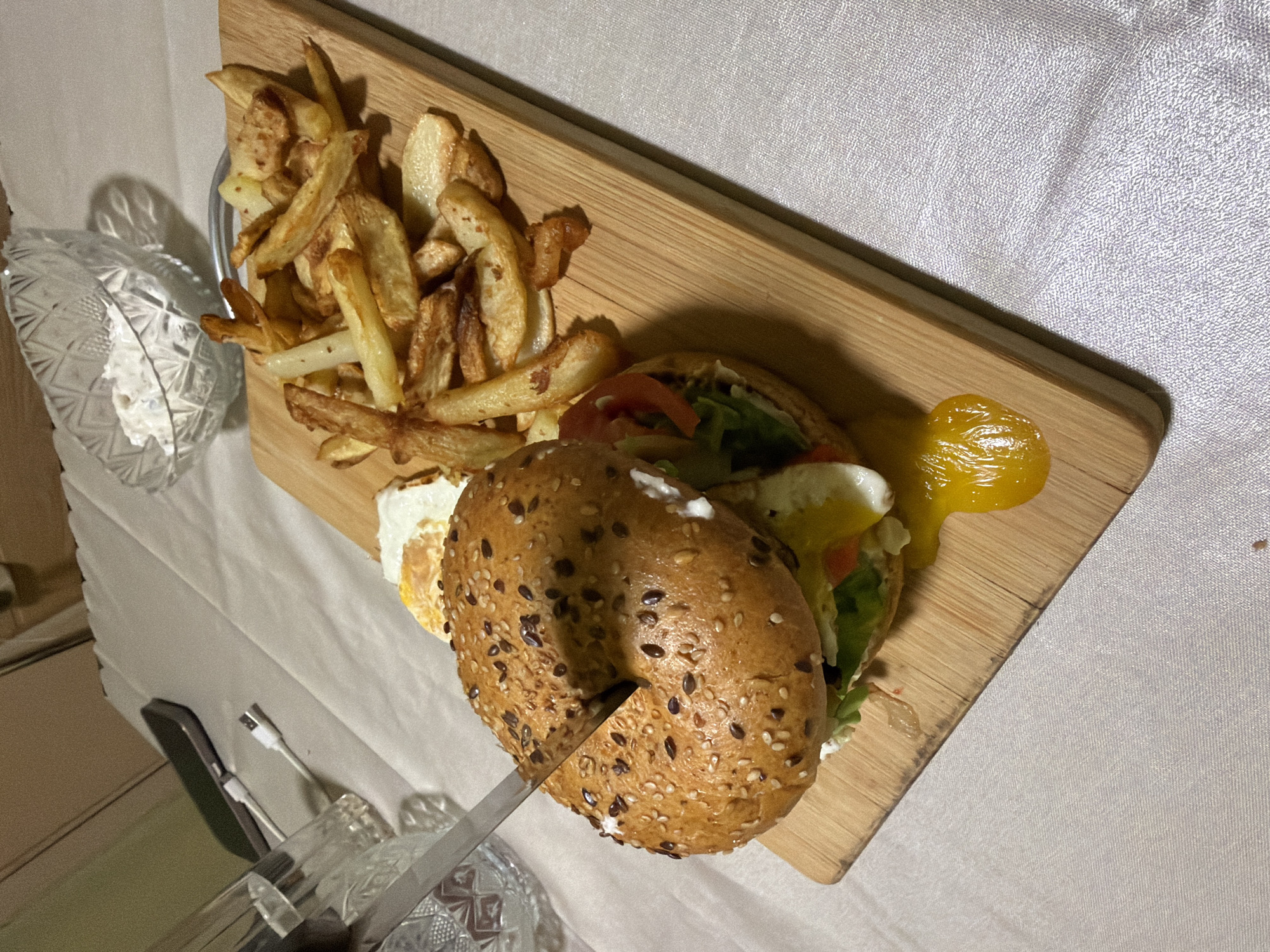 The image depicts a wooden cutting board with food on a textured white surface, likely a tablecloth. On the board, there is a pile of fried potatoes, a cut bagel sandwich with visible lettuce, tomato, egg, and cream cheese, and a dollop of yellow sauce. A knife is cutting the bagel. The scene includes a clear, patterned glass bowl and a cutlery set partially visible in the lower left corner. The environment suggests an indoor dining setting. There is no indication in the image of the location being Varna, Bulgaria.