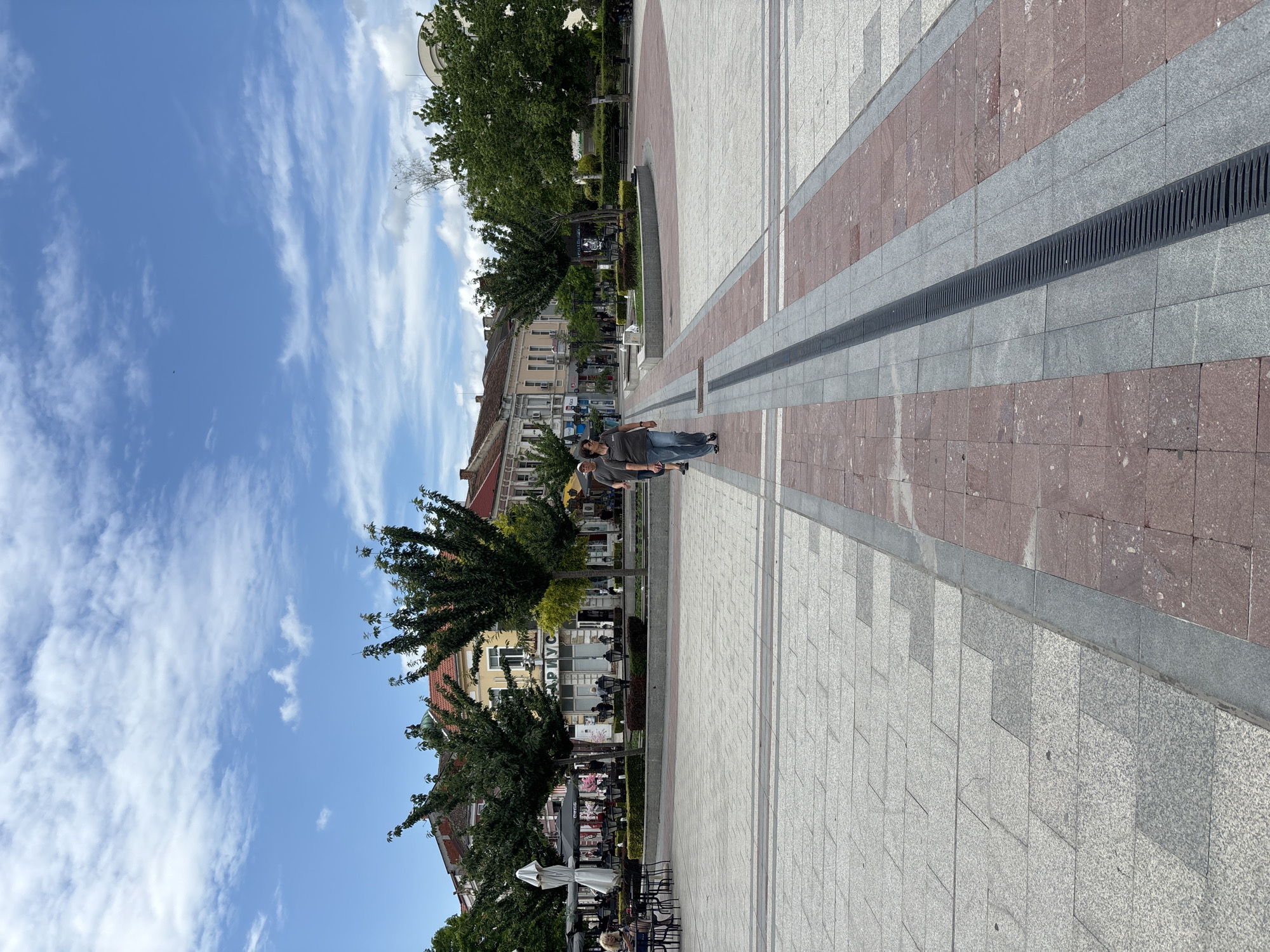 The image, rotated 90 degrees clockwise, presents a bright public square or pedestrian area under a partly cloudy, blue sky. The ground is paved with a distinctive pattern of light grey, dark grey, and reddish-brown rectangular tiles. A prominent, long black gridded drainage channel runs down the center of the main pathway.
In the middle ground, two individuals are walking on the paved surface; one is clearly visible wearing a grey t-shirt and jeans, while the other is partially obscured. Further in the background, several people are seated at outdoor tables beneath umbrellas, indicating the presence of cafes or restaurants.
The square is bordered by multi-story buildings, showcasing diverse architectural styles, some featuring reddish-brown tiled roofs. Numerous lush green trees, some planted in decorative beds, line the pedestrian zone. On a yellow building in the background, Cyrillic text is visible above an awning, spelling "АРИУС" (ARIUS).
The scene suggests a sunny late morning or early afternoon, with clear skies and scattered white clouds. The atmosphere is calm and leisurely, depicting typical daily activity in what appears to be a European urban setting, potentially in a Slavic-speaking country due to the visible text.