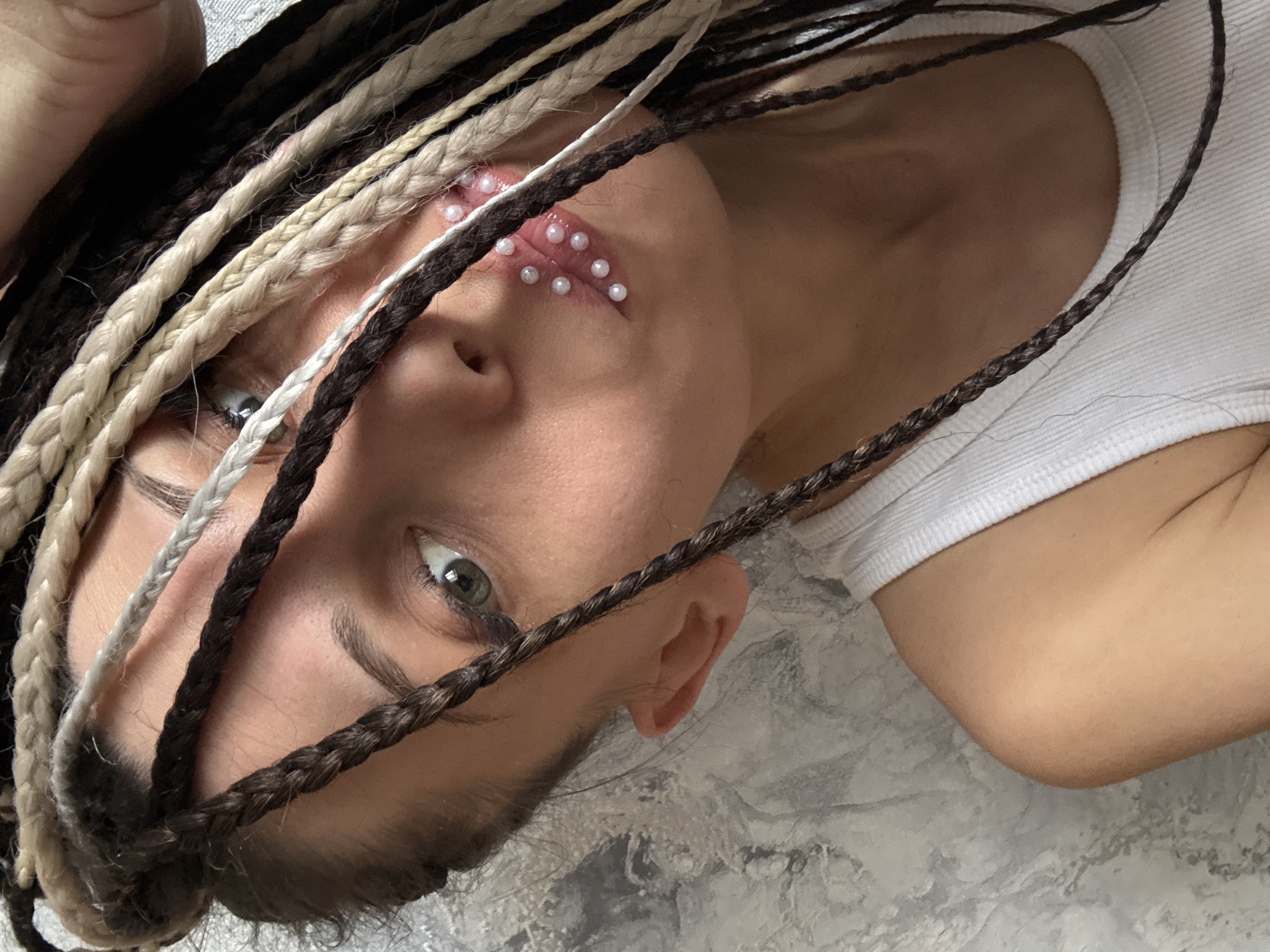 The image presents an artistic, close-up, and angled portrait of a young person, likely female, set in Rusanovo, Russia. The subject is shown from the chest up, with her head tilted, and she is looking directly at the viewer with her left eye (from her perspective).
Her hair is styled in numerous multi-colored dreadlocks or braids, featuring shades of dark brown, light brown/beige, and white/grey, which frame her face and cascade across her chest. A prominent and unique detail is the adornment on and around her lips: several small, lustrous white pearl-like beads are meticulously arranged in a pattern across her upper and lower lips. Her visible eye is a striking light green or hazel.
She is wearing a simple white ribbed tank top or undershirt. The background appears to be a light gray or white textured surface, possibly a wall or bedding, providing a neutral backdrop that highlights the subject. The lighting is soft and even, creating a gentle illumination without harsh shadows. Her expression is calm and direct. There is no visible text in the image.