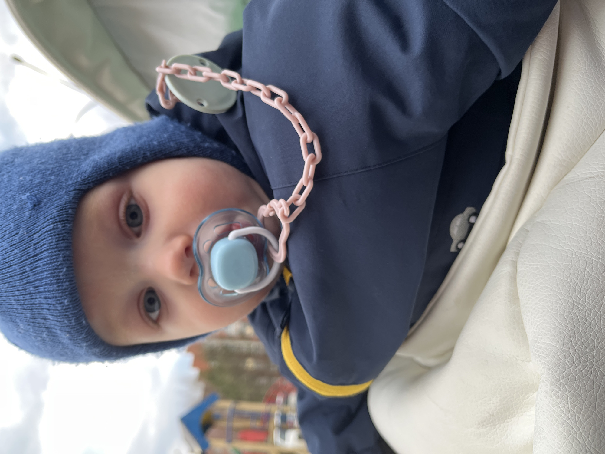 A close-up photograph, rotated sideways, captures a baby in a stroller looking directly at the camera. The baby has bright blue eyes and is dressed for cool weather in a blue knitted hat and a dark blue padded jacket featuring a narrow yellow stripe on the sleeve. A light blue pacifier is held in the baby's mouth, connected by a pink plastic chain to a light green circular clip fastened to the jacket. The baby's skin appears fair. To the right, part of the stroller's interior is visible, made of a white, textured material resembling leather. The background is out of focus, showing a bright sky and distant, blurry shapes of colorful playground equipment (blue, red, yellow), suggesting an outdoor setting like a park in Chaikovsky, Russia. The clear lighting indicates it is daytime. The baby's expression is neutral and curious. No text is visible in the image.