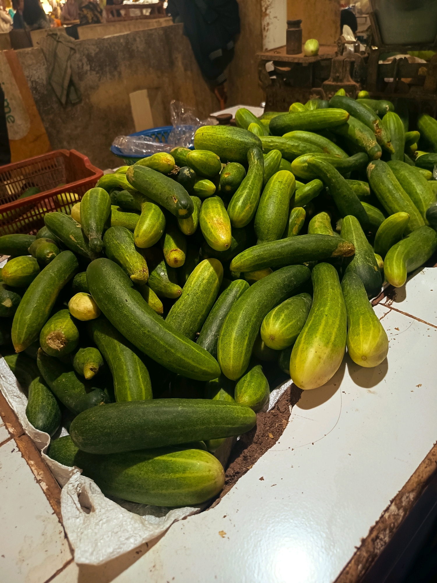 A pile of fresh cucumbers sits on a white table, possibly at a market in Batang, Indonesia. The cucumbers are mostly green with some showing yellowing, suggesting ripeness. Some are smooth and elongated, while others are more bulbous. A red plastic basket containing more cucumbers is visible to the left. In the background, out of focus, are hints of market stalls, weighing scales, and people. The lighting suggests an indoor setting, possibly during the day or under artificial light.