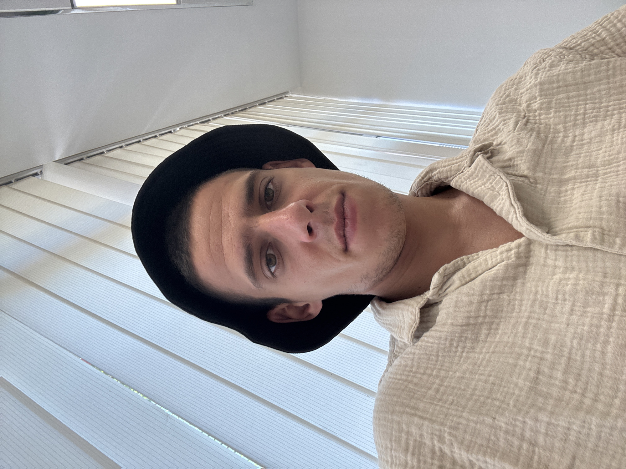 A young man with short dark hair, wearing a black bucket hat and a light-colored, crinkled, button-down shirt, is pictured in a selfie. He is looking directly at the camera with a neutral expression. The background consists of vertical blinds, suggesting an indoor setting, possibly an office or a room with windows. The lighting appears to be natural, coming from the blinds. The image has a clean and simple aesthetic, with the focus solely on the subject. This scene could be located in any urban environment; no specific location in Sofia, Bulgaria, can be determined from the visual information provided.