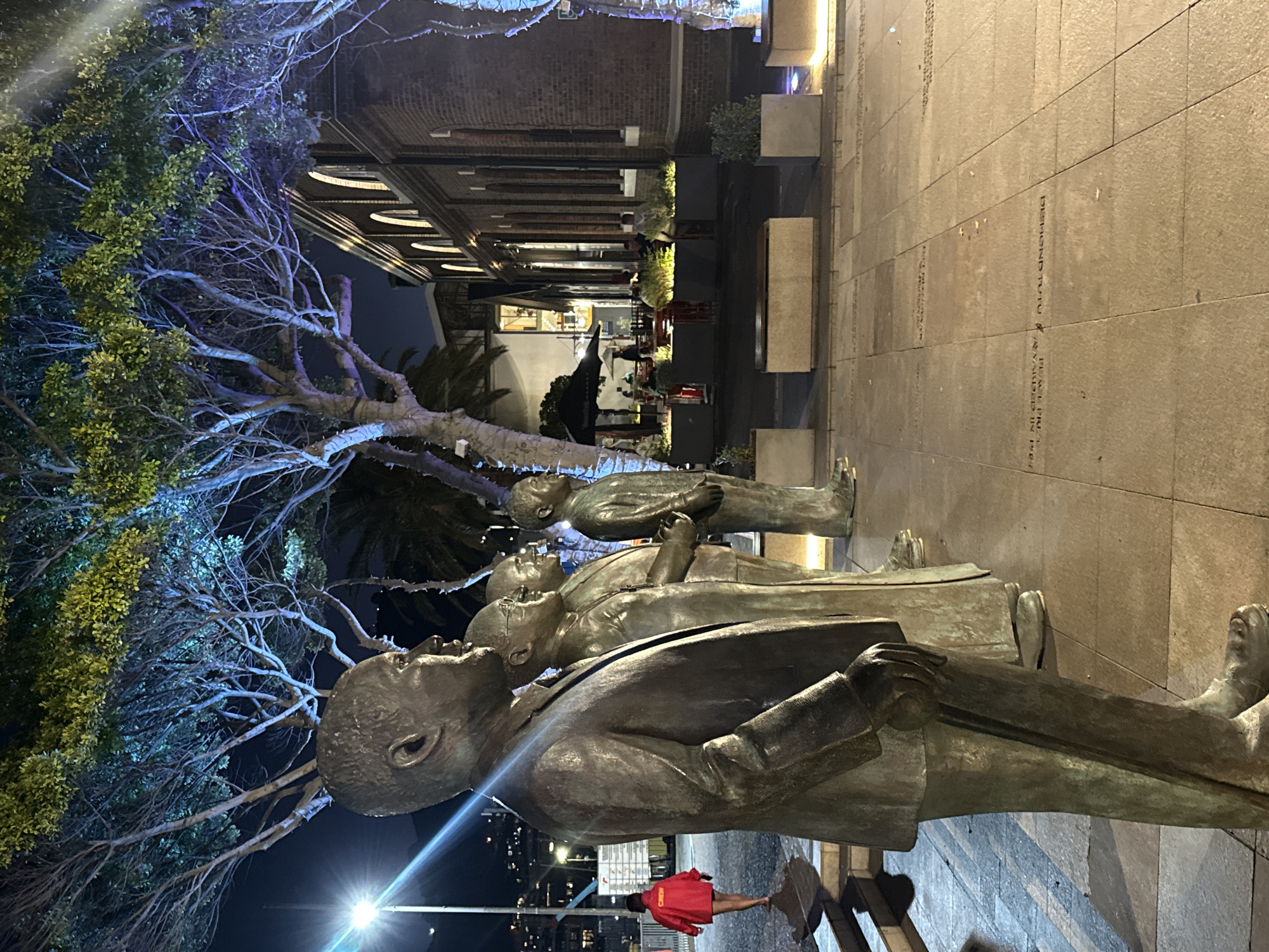 The photograph depicts a bronze statue of several figures in business suits, likely depicting an important moment of people leading to the end of apartheid. The statue is located on a paved walkway in Cape Town, South Africa, at night, with trees on the left side of the image lit with blue light. A building can be seen in the background, with trees and other greenery, and other figures walking in the distance. A person wearing a red jacket can be seen near the base of the statue on the right. The scene is illuminated by streetlights, and the overall tone is subdued.