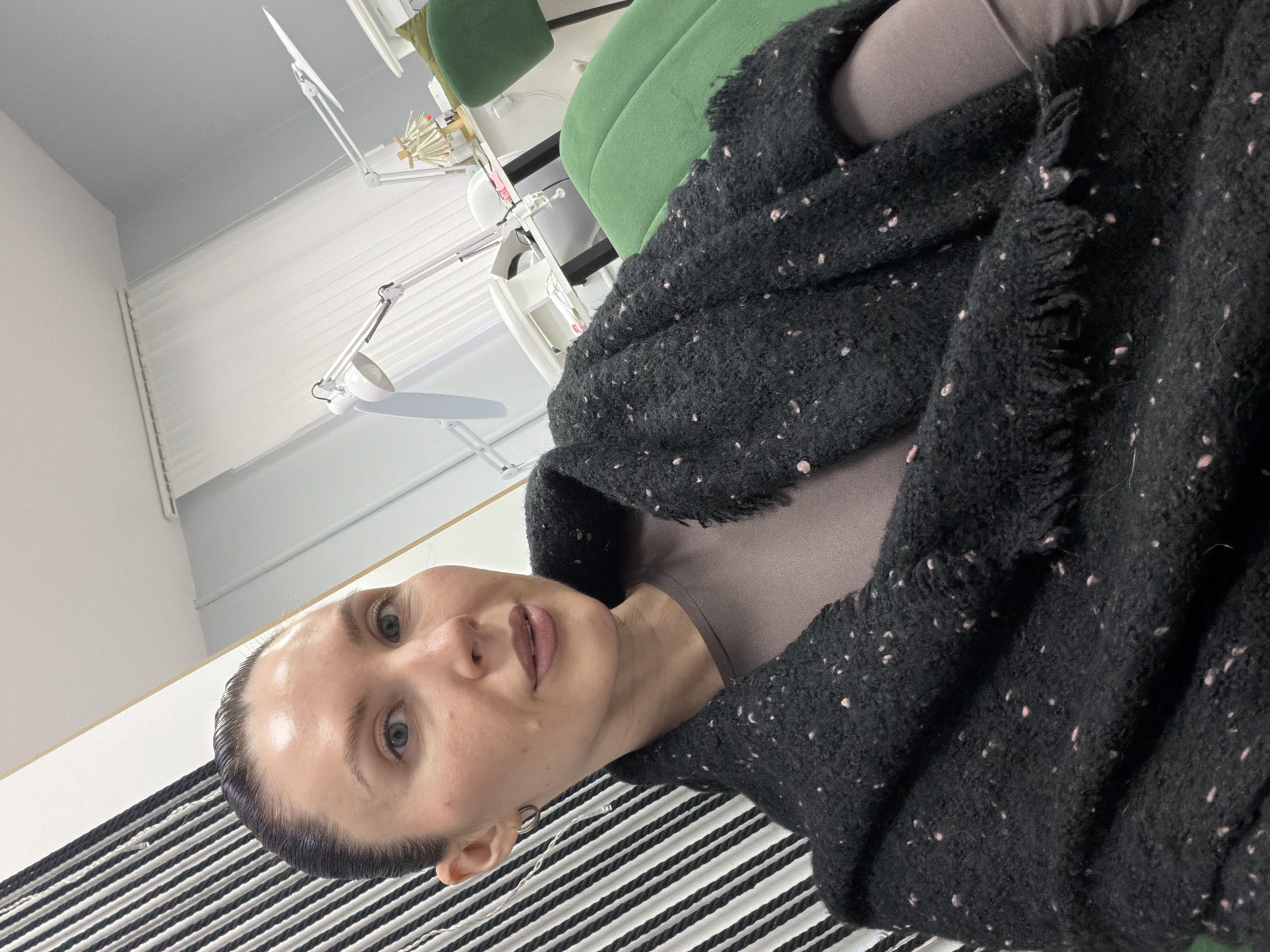 A woman with short, dark hair pulled back from her face is indoors, wearing a heathered gray shirt and a large, dark gray shawl or scarf with pinkish flecks. She is looking toward the camera. Her left ear is visible, pierced with a silver hoop earring. The background includes a green chair and a table with white task lamps. To the left, there is a white wall with a row of narrow windows. The woman is sitting on a black and white striped surface, possibly a cushion or a piece of furniture. The photograph is taken from a slightly low angle and appears to be rotated counterclockwise. This scene could be in a salon or a medical office in Perm, Russia.