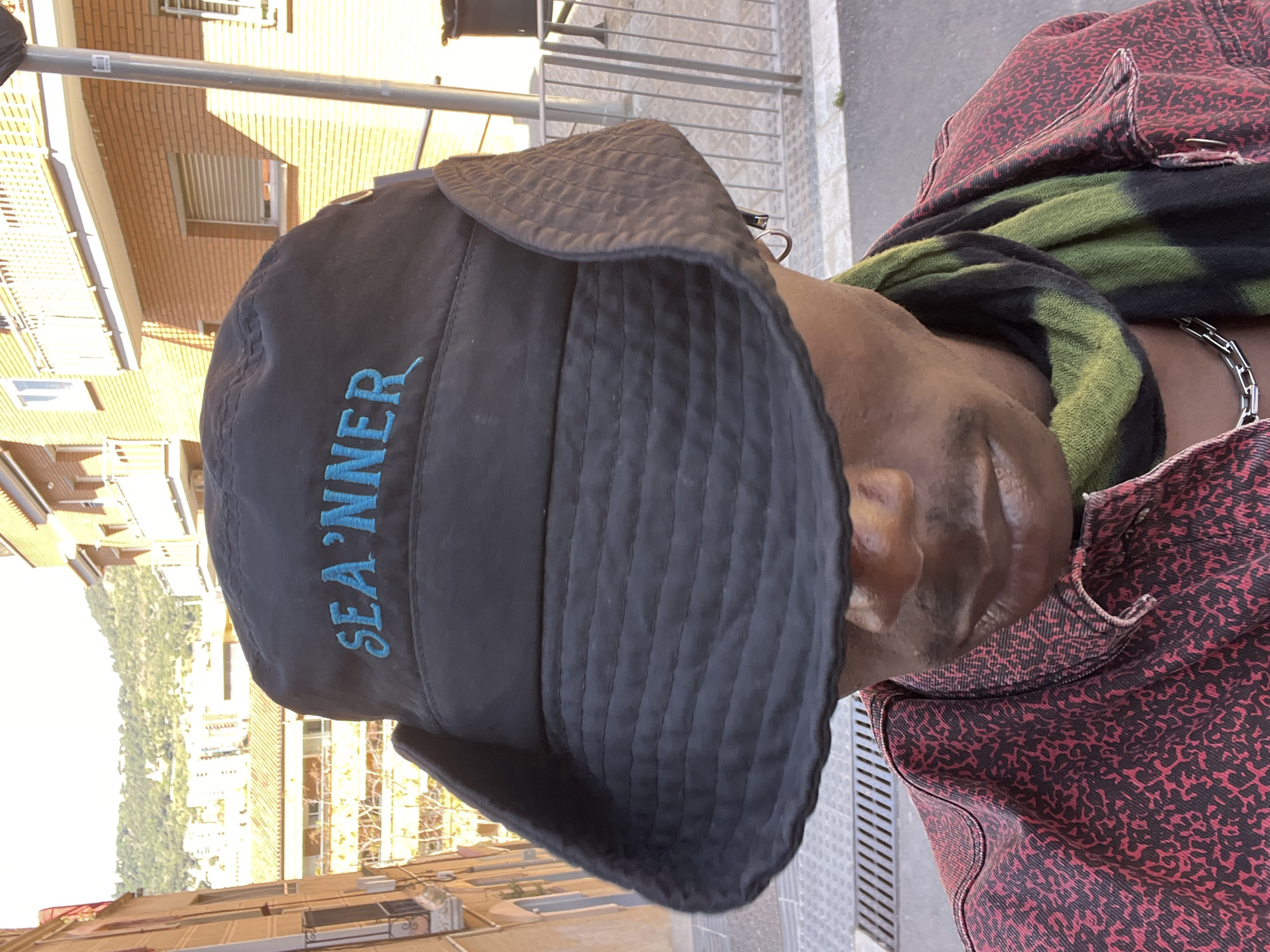 A sideways portrait captures a person from the chest up, focusing on their attire and partial face in an urban outdoor setting, consistent with Barcelona, Spain. The subject has dark skin, with only the lower part of their face (jawline, ear, cheek, and mouth area) visible. They are wearing a black bucket hat with "SEA'NNER" embroidered in light blue on the front. A green and black tie-dye scarf or similar garment is wrapped around their neck, partially covered by a red and black patterned jacket, which appears to be made of denim or a sturdy fabric. A silver chain necklace is visible around their neck.
The background shows brick-faced buildings under bright daylight, suggesting a sunny day, with strong shadows indicating either morning or afternoon sun. Metal railings are also visible in the background, possibly part of a balcony or street barrier. The person's expression appears neutral to slightly smiling.