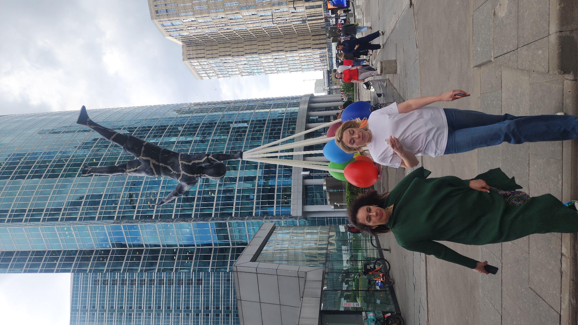 The image shows two women standing in front of the skyscraper in the Moscow International Business Center, also known as Moscow City. One woman is wearing a white t-shirt and jeans, while the other is wearing a green overcoat. They are holding balloons with string that is attached to a large statue of a diver on the side of the building. There are other people walking by in the background.