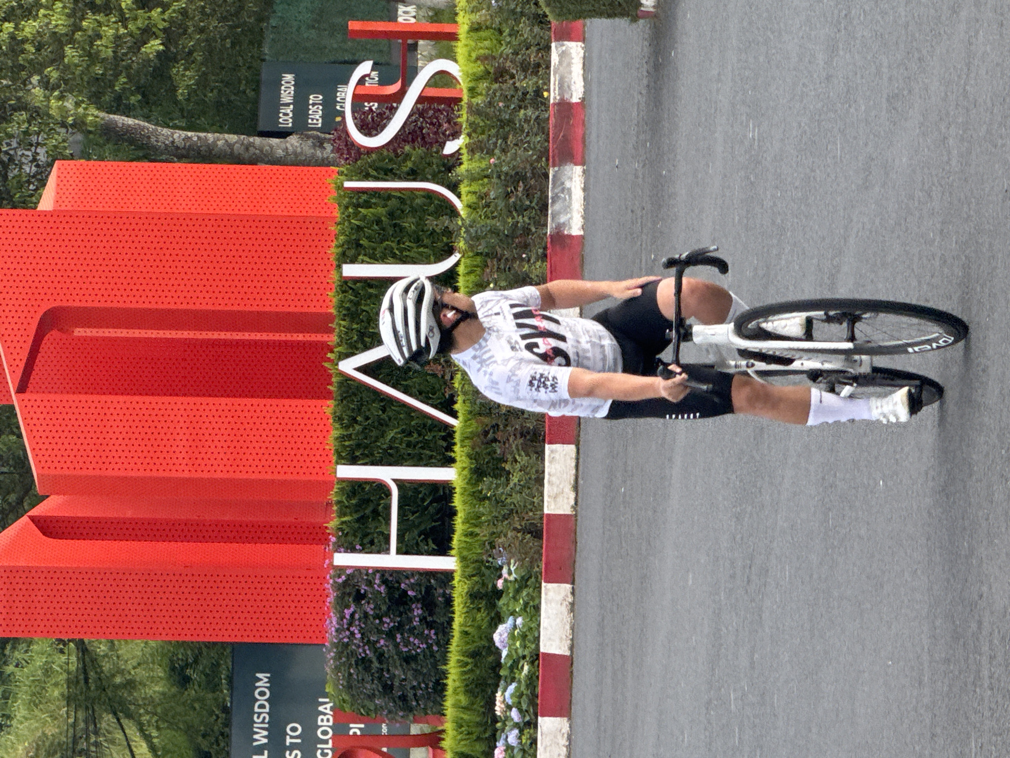A person is riding a bicycle on a road next to a large, red, modern art sculpture and a manicured hedge. The person is wearing a white and black cycling helmet, a white t-shirt with black graphics, black cycling shorts, and white socks. They are also wearing cycling shoes. The bicycle is white with black tires. The sculpture is made of bright red, perforated metal panels, arranged in a way that resembles stacked boxes. The hedge is green and well-maintained, with some flowering plants in shades of pink and blue at its base. A sidewalk with red and white striped borders is visible between the road and the hedge.
In the background, there are trees and a dark sign with white text that reads "LOCAL WISDOM LEADS TO GLOBAL ACTION". The scene appears to be outdoors during the day. The weather is overcast, judging by the diffused light and the slightly wet appearance of the road, with some faint streaks that might indicate light rain or water droplets on the lens. The overall atmosphere is one of an athletic activity taking place in an urban or park-like setting with artistic elements. The text suggests an educational or philosophical context for the location.