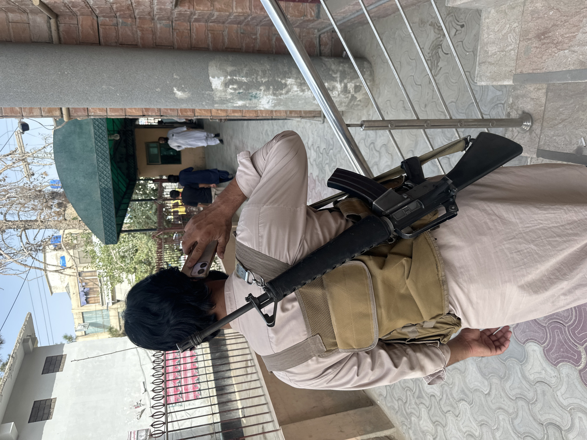 The image shows a man from behind, walking on a paved pathway in Quetta City Tehsil, Pakistan. He is wearing a light-colored tunic and trousers, a tactical vest, and has a rifle slung over his right shoulder, with its barrel pointing upwards. The rifle appears to be an AR-15 style weapon. The man is holding a mobile phone to his right ear with his right hand, engaged in a phone call. He is walking away from the camera, towards an entrance with a green awning. In the background, other people can be seen walking, and there are buildings with various architectural styles. The environment suggests an outdoor setting in a moderately urban area.