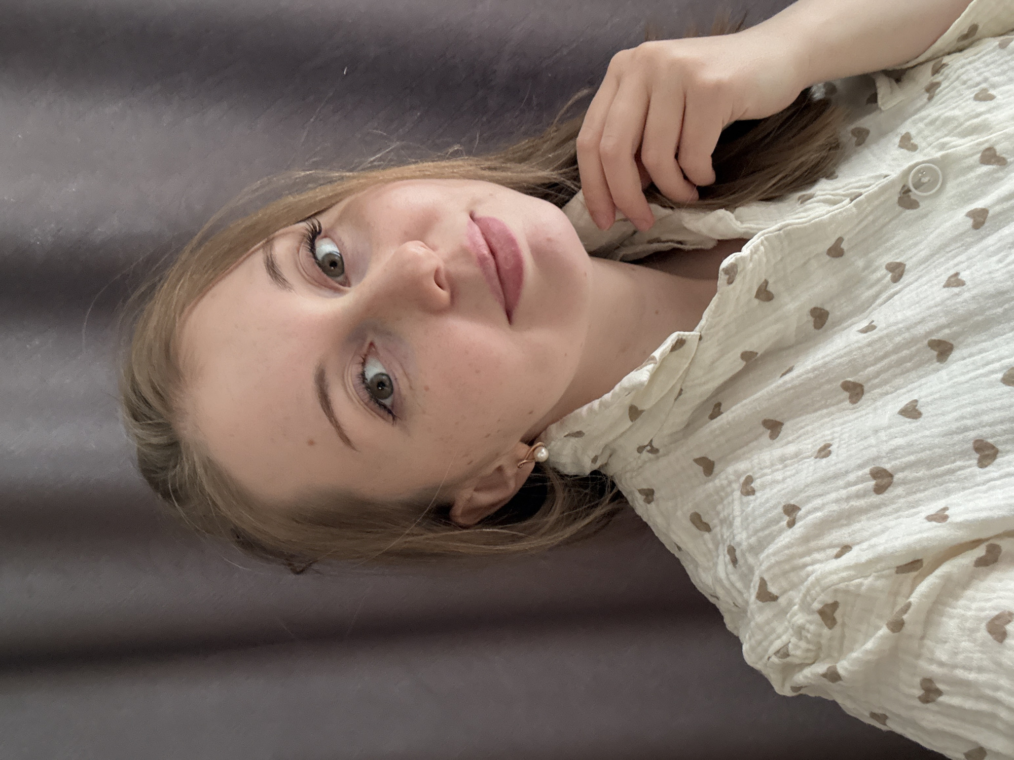 A young woman is captured in a close-up portrait, positioned horizontally across the frame with her head tilted. She has fair skin, visible freckles, and light brown hair. Her green eyes are open, looking directly at the viewer with a calm and gentle expression. Her lips are slightly parted and appear to have a soft pink tint. She is wearing a light-colored, possibly cream or off-white, collared shirt patterned with small brown hearts. A white button is visible on the shirt. A delicate pearl stud earring, adorned with a thin rose gold-colored wire, is visible in her right ear. Her right hand is resting near her head, touching her hair. The background is a plain, dark gray or brown fabric, suggesting an indoor setting. The lighting is soft and even, highlighting her features without harsh shadows. No text is visible.
