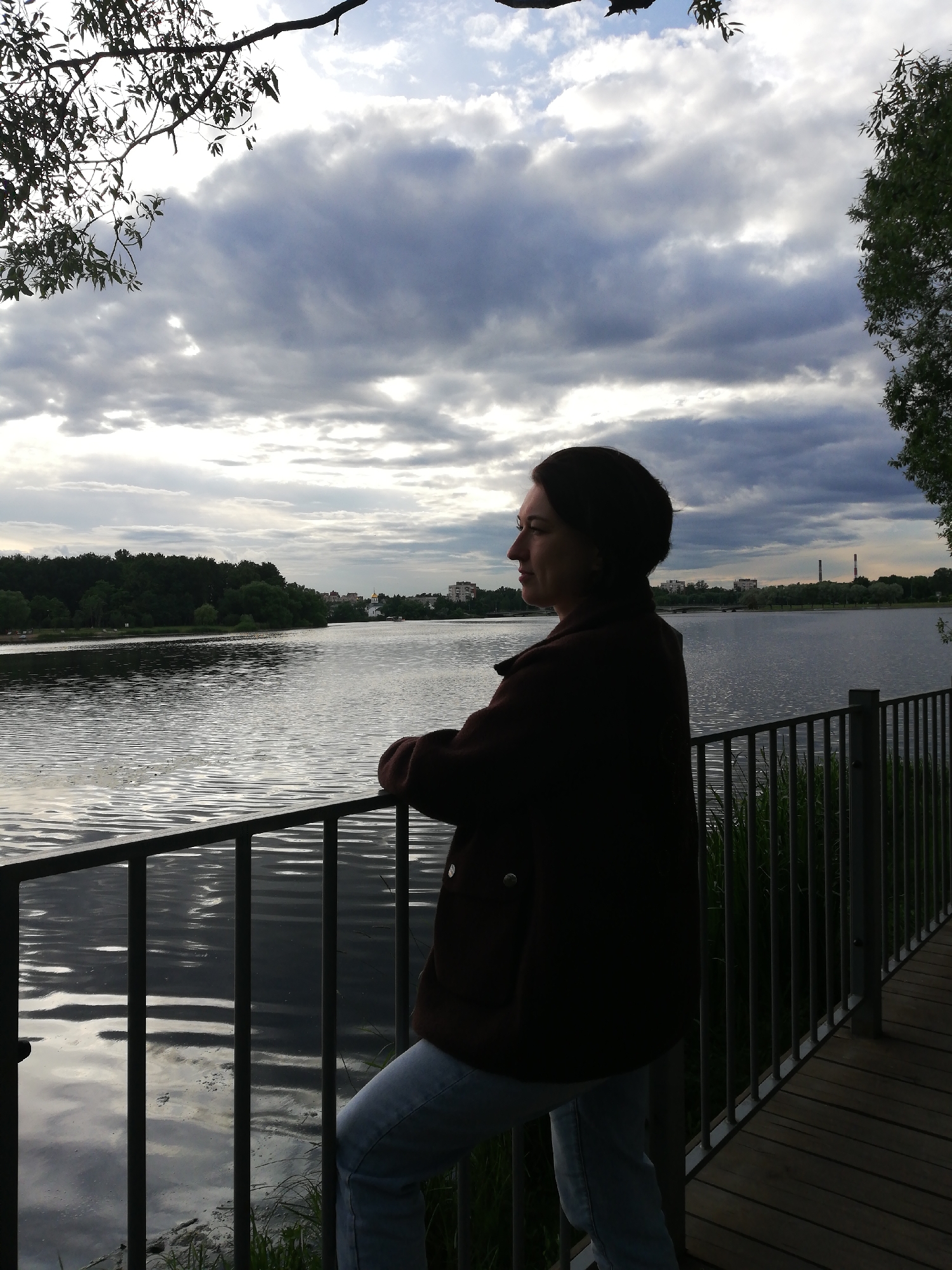 A woman stands with her back to the camera, looking out at a body of water. She is wearing a dark jacket and light-colored pants. A metal railing is in front of her, and a wooden walkway is to her right. The water reflects the cloudy sky. Trees line the far bank of the water, with some buildings visible in the distance. The sky is full of dramatic, grey clouds, suggesting it might be overcast or preparing to rain. The light is diffused, indicating it is likely daytime, possibly late afternoon or early evening. The overall mood is contemplative, with the woman appearing to be lost in thought as she gazes at the landscape. No text is visible in the image.