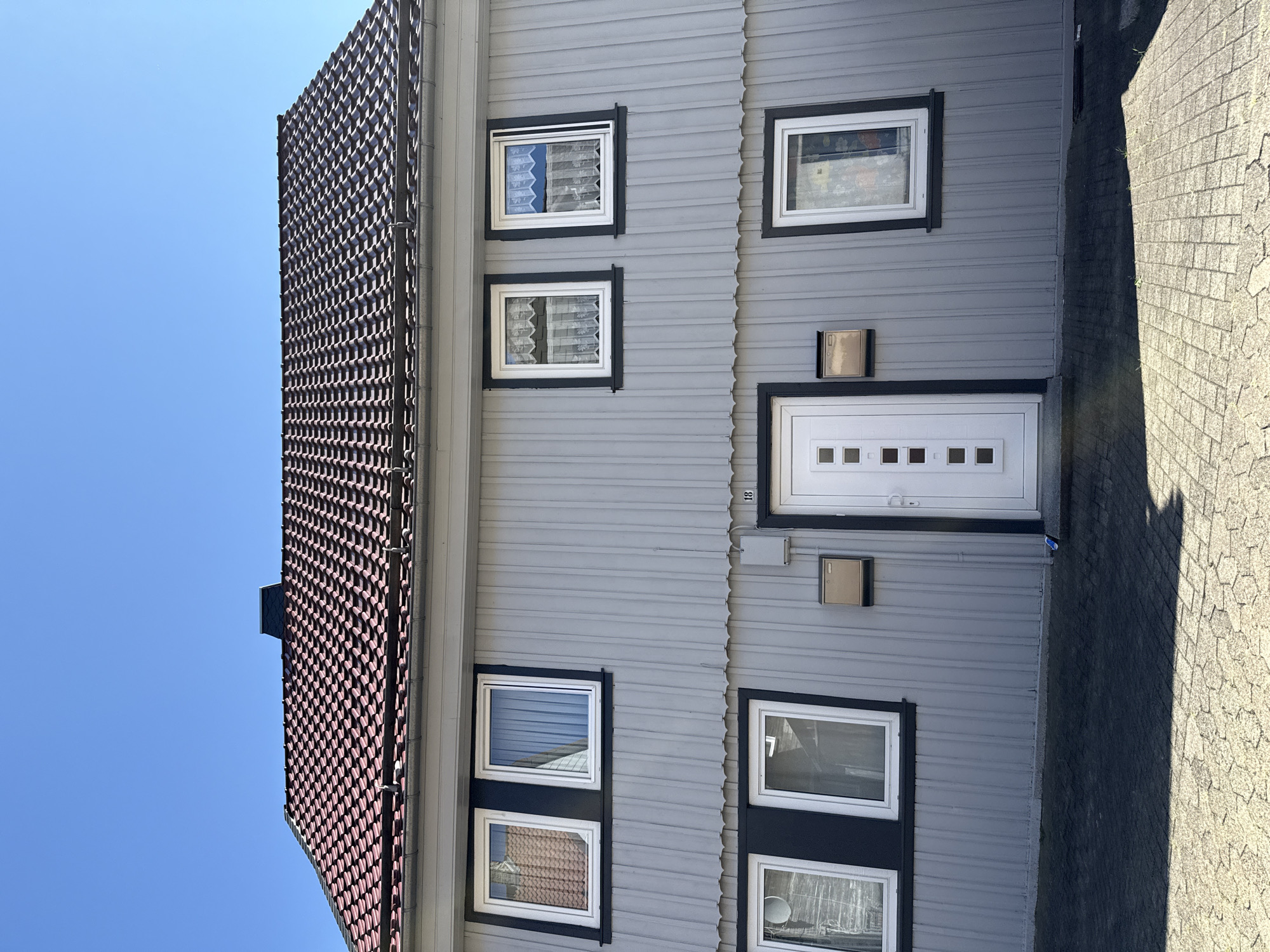 The image shows the front of a two-story, light gray, wood-paneled house under a clear blue sky. The house has a dark red tiled roof and a black chimney. There are multiple windows with white frames and dark trim on both floors. The upper floor has two windows on the left, one reflecting the red roof, and two windows on the right, both with white lace curtains. The lower floor has two windows on the left and one on the right, all with white frames and dark trim. A white front door with square glass panels is in the center of the lower floor, flanked by two mailboxes. The house number "18" is visible above the door. The ground in front of the house is paved with hexagonal interlocking stones, with a dark shadow cast across it. The scene appears to be in Herzberg, Germany.