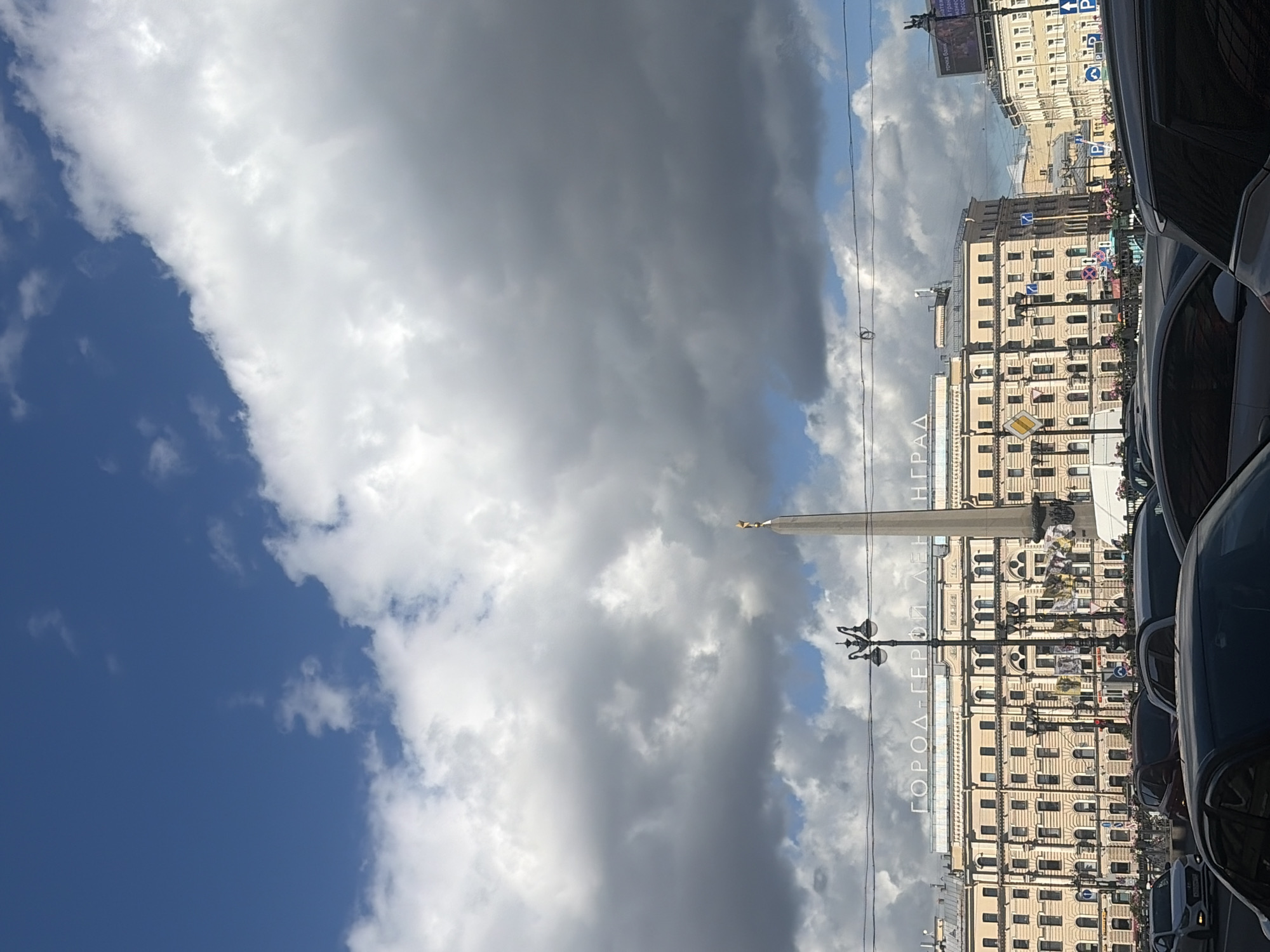 The image captures a wide-angle view of a street in Saint Petersburg, Russia, on a cloudy day. In the foreground, several cars are visible, suggesting traffic on the road. Behind the cars, a large, light-colored building dominates the scene, with multiple windows and architectural details. An obelisk, likely a monument, stands tall next to the building, topped with a golden figure. The sky above is filled with dramatic, dark clouds interspersed with patches of blue. A street lamp is positioned in front of the obelisk, adding to the urban landscape. The text "ГОРОД-ГЕРОЙ ЛЕНИНГРАД" (Hero City Leningrad) is visible in large letters across the facade of the building, indicating the location's historical significance. Further in the background, more buildings and street elements contribute to the dense urban environment.