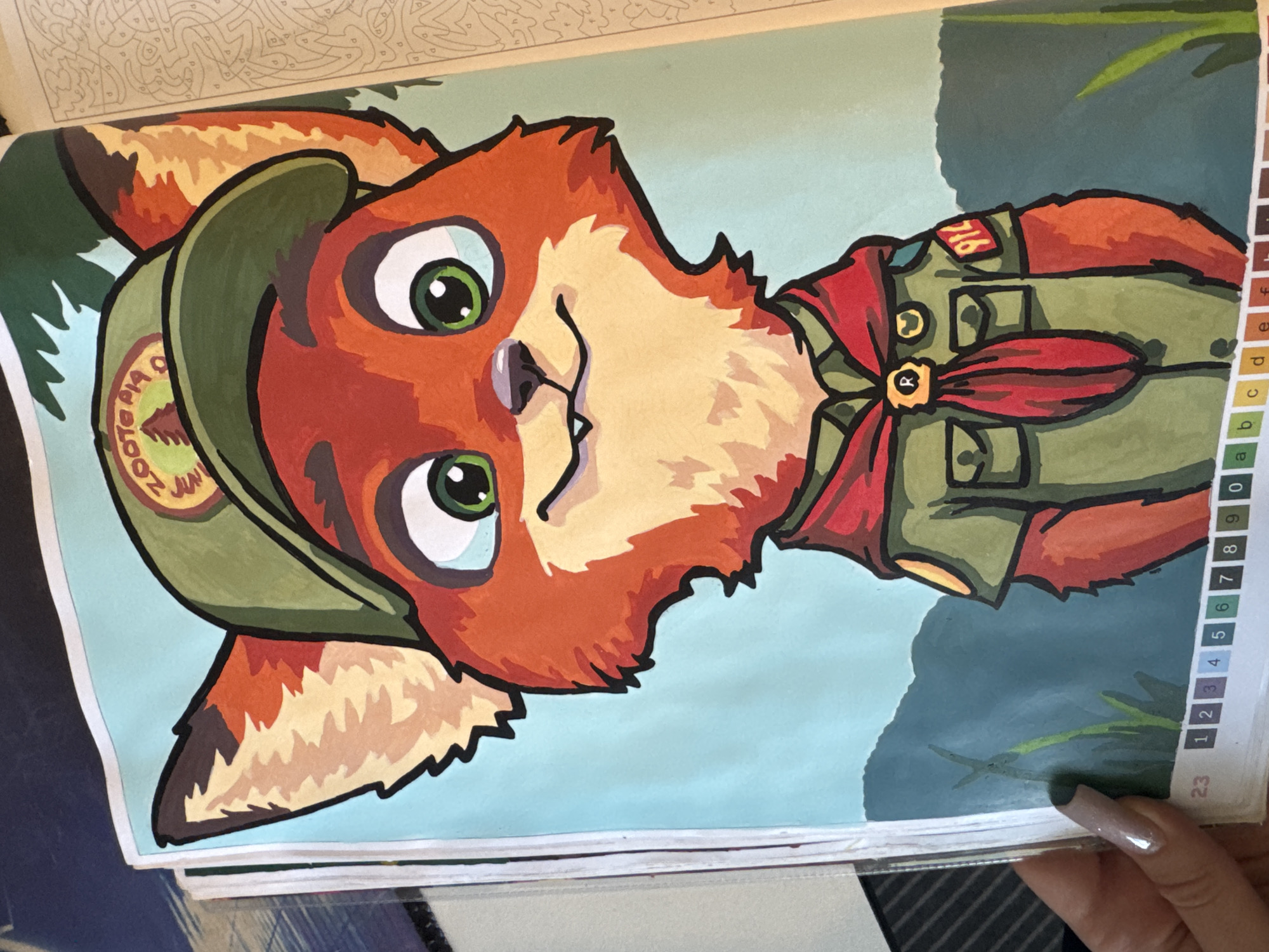 A painting of a cartoon fox character, dressed as a scout, is displayed on a page. The fox has orange fur, green eyes, and is wearing a green scout hat with a patch that reads "ZOOTOPIA". It also wears a green uniform with a red scarf, a badge with an "R", and another badge with the number "914". The background consists of a light blue sky and green, silhouetted hills. The bottom edge of the page shows a numbered color key. The image is cropped, with a hand visible on the left side.