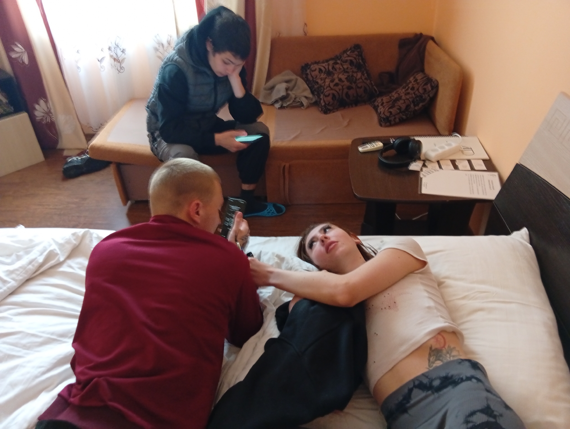 The image depicts an indoor scene. A woman lies on a bed, while a man in a red shirt is next to her, looking at a phone he is holding. A third person is sitting on a couch in the background, looking at their own phone. The couch has pillows on it. A small table next to the couch holds items including headphones, a remote, and papers. A tattoo is visible on the woman's side. The location is possibly a room within a building in the city of Poltava, Ukraine, based on additional context provided by the user.