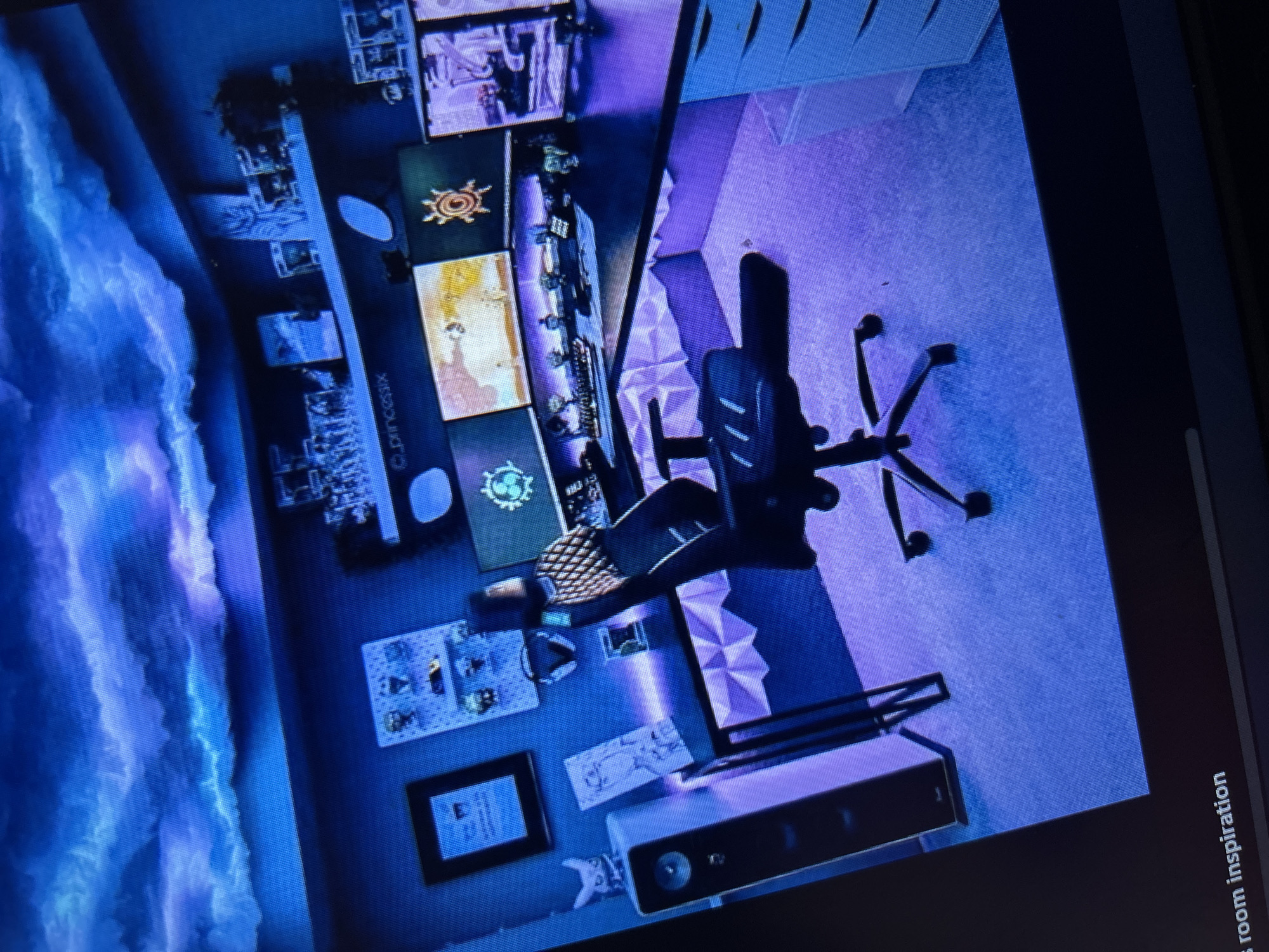 The image shows a gaming room. The room features a large wall-mounted screen displaying an image of clouds. There are multiple smaller screens arranged in a vertical stack, displaying various images and graphics. A black gaming chair is centrally positioned, facing the screens. Decorative panels are visible on the wall next to the desk. A speaker or soundbar is located on a surface near the bottom of the image. Artwork, collectibles, and a framed picture are also present. The room is illuminated with a predominantly blue and purple lighting scheme. The words "room inspiration" are present in the bottom right corner. The image does not contain enough information to determine the location in Sofia, Bulgaria.