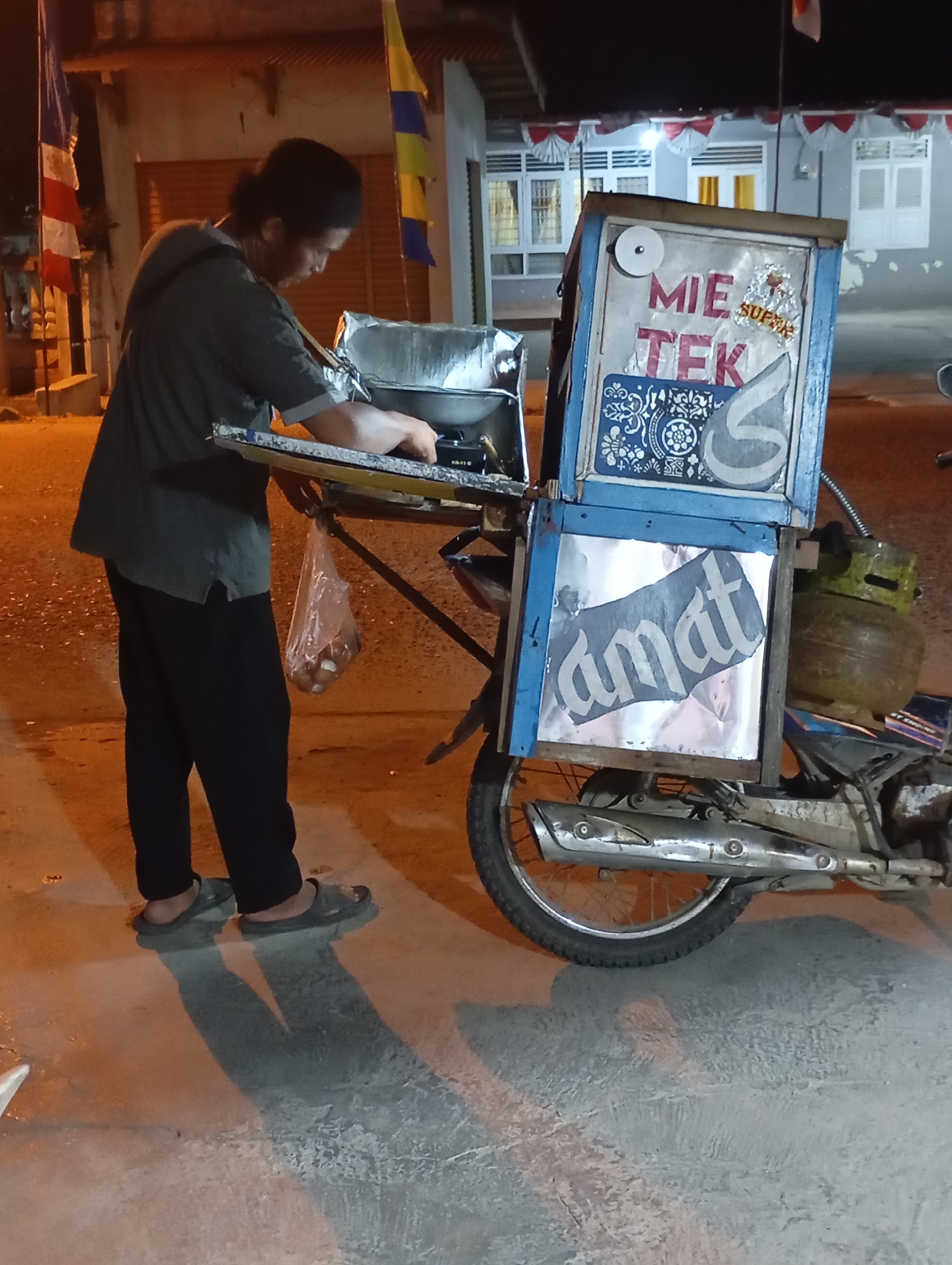A person, viewed from the side, is preparing food at a mobile cart attached to a motorcycle. The cart is blue and has signs with "MIE TEK" and "amat" written on it. The person is wearing a dark green shirt and black pants. A plastic bag containing food hangs from the cart. The scene appears to be a street at night, with lights illuminating the area and casting shadows. In the background, there are buildings and flags, suggesting an urban setting in Metro, Indonesia.