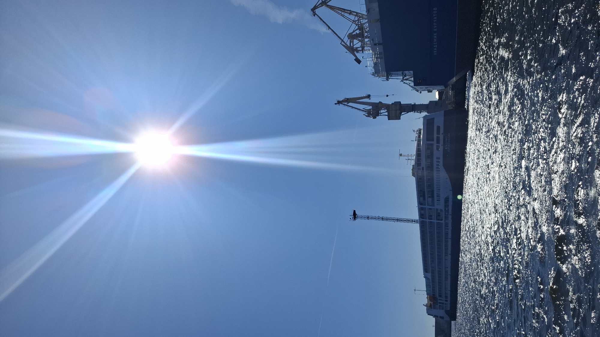 A brightly sunlit scene shows a body of water with ships docked at a port. The sun is high in the sky, casting bright rays downwards. In the foreground, the water is choppy and reflects the sunlight. To the left, a large, white passenger ship is docked. It has multiple decks and windows. To the right, a large blue ship is docked, with the text "АКАДЕМИК КУРЧАТОВ" (Academic Kurchatov) visible on its side. Several cranes are also present in the port, suggesting ongoing industrial activity. The sky is clear and blue, with a faint contrail visible in the distance. The location is likely a port in Saint Petersburg, Russia, possibly the Passenger Port of St. Petersburg (Marine Facade). There are no people visible in the frame.