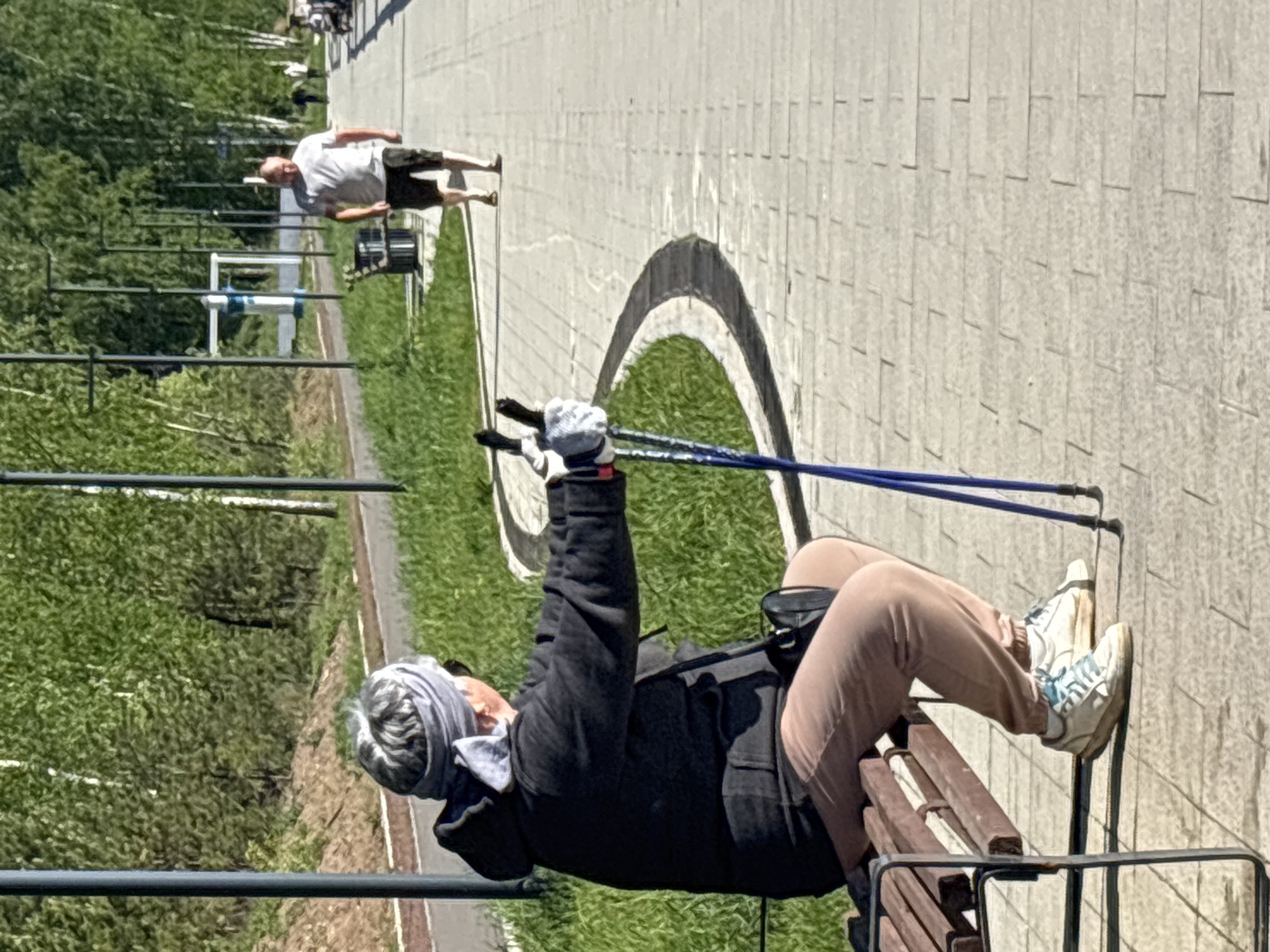 An outdoor scene in Chaikovsky, Russia shows two people. In the foreground, a woman with short gray hair covered by a light gray headscarf sits on a wooden bench. She wears a dark jacket, light brown pants, and white sneakers with blue accents. She holds the handles of two blue Nordic walking poles. Her legs are extended, and her feet are resting on the bench's backrest. In the background, a man in a white t-shirt and dark shorts walks along a sidewalk paved with light gray tiles. A grass area with fitness equipment runs parallel to the sidewalk, separated by a metal fence. A dark arch-shaped design is marked on the pavement. The lighting suggests a sunny day.