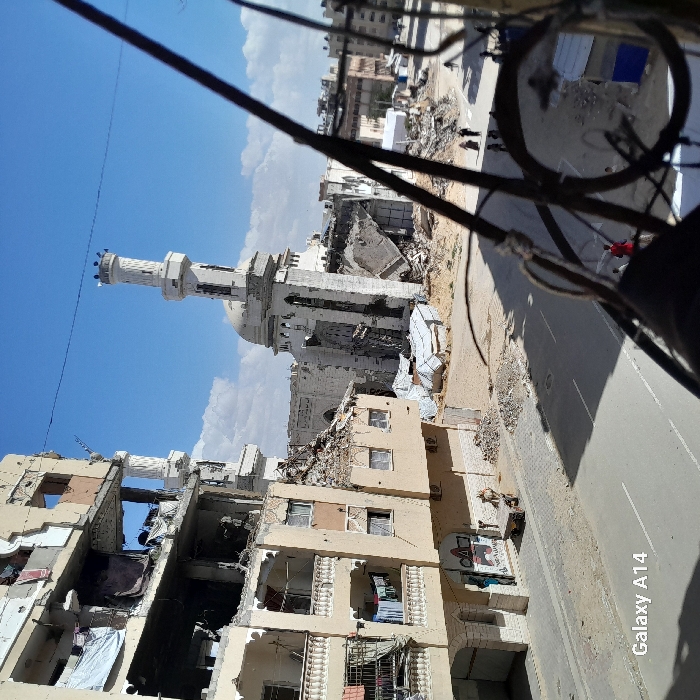 The image captures an urban scene of extensive destruction in Khan Yunis, Palestinian Territory, under a partly cloudy blue sky. The elevated perspective overlooks a street flanked by severely damaged structures.
To the immediate left, a multi-story residential building stands heavily compromised, displaying multiple breached exterior walls that expose internal spaces, fractured concrete, and exposed rebar. White sheets are draped over several upper-level openings, and articles of laundry hang from a balcony railing on a lower floor. The ground floor features a commercial facade with a visible "ΔΨ" symbol. On the sidewalk below, a male figure in light-colored attire stands next to a two-wheeled utility cart.
A paved road spans the midground, appearing clear of major debris but bordered by rubble and dirt. In the midground and extending into the background, a large mosque, characterized by its white dome and two minarets, has sustained catastrophic damage. The main prayer hall and significant portions of its structure are collapsed into a massive pile of concrete and debris. One minaret remains largely upright but exhibits visible structural compromise, while the base of the other is part of the collapsed structure. Extensive piles of construction rubble and several makeshift shelters constructed from tarpaulins surround the mosque.
In the distant background, additional urban buildings are visible. On the far side of the street, approximately 5-7 indistinct figures are observed walking. A solitary figure wearing a red garment is partially visible closer to the right edge of the frame. The extreme right foreground is partially obscured by out-of-focus, dark metal components, suggesting the photograph was taken from behind a railing or similar barrier. A "Galaxy A14" watermark is present in the bottom-left corner.