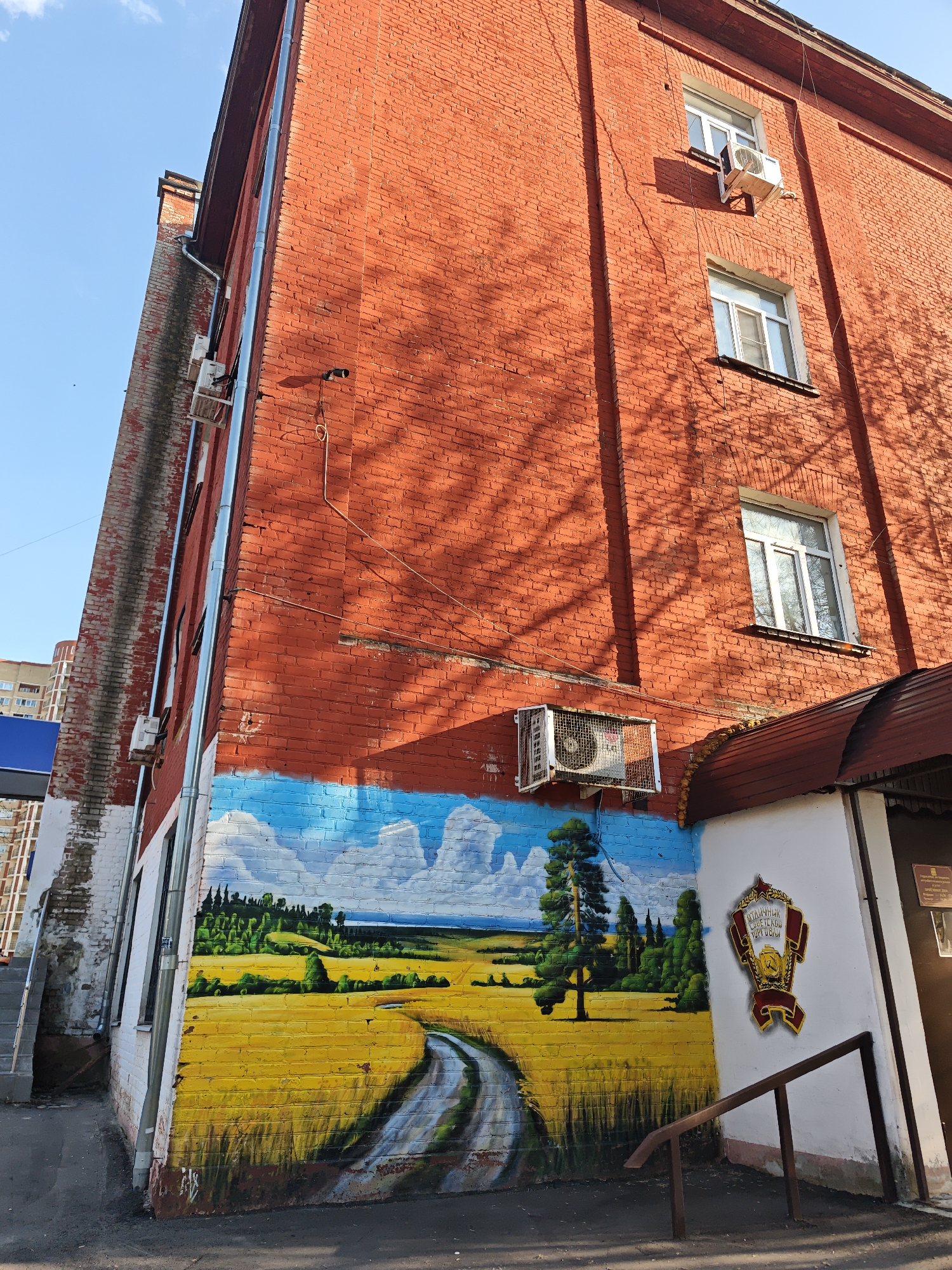 The exterior corner of a three-story brick building is visible under a partly cloudy sky in the city of Unknown, Unknown. The building is primarily red brick, with a mural painted on the lower corner section. The mural depicts a blue sky with white clouds transitioning into a landscape of green trees and a yellow field with a winding gray path.
On the side of the building with the entrance, next to the mural, there is a white-painted area with a decorative emblem featuring a hammer and sickle. The emblem is red, gold, and white. A brown handrail leads up to the entrance.
There are several windows visible on the brick building, along with air conditioning units mounted on the exterior. The overall impression is of an older building with recent additions and artistic decoration. A dark gray sidewalk extends along the base of the building.