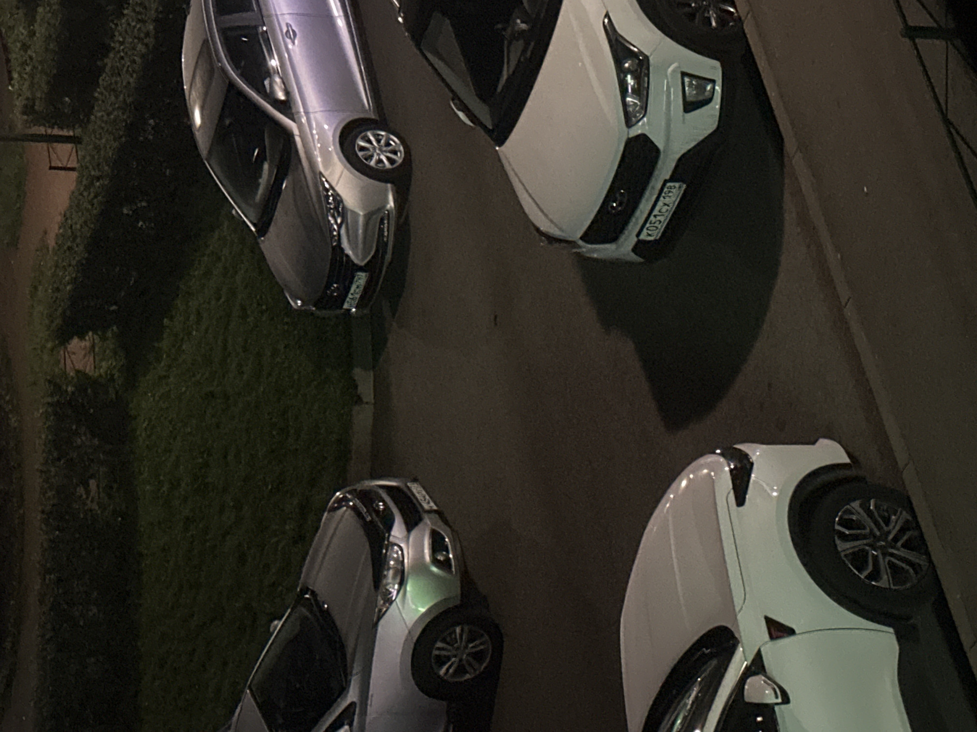 The image shows a nighttime view of a parking area with four cars. Two silver cars and two white cars are visible. One silver car is positioned on the left, partially obscured by a hedge. Another silver car is on the upper right. Two white cars are on the lower right and lower left of the frame. The car on the lower right has a license plate that reads "K051Cx 198". The ground is paved asphalt, and there is a curb in the foreground. Green hedges and grass are visible behind the cars. The lighting suggests it is nighttime.