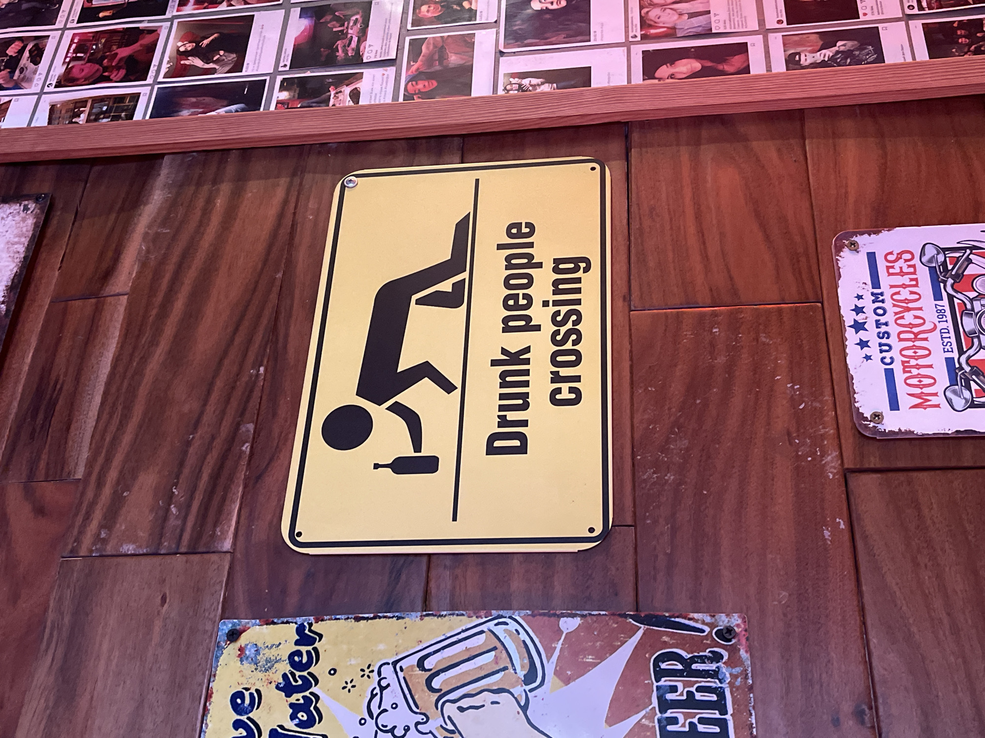 The scene depicts a close-up view of a wooden wall with various signs attached to it. In the center is a yellow rectangular sign with a black graphic of a person falling over, presumably due to intoxication, along with the words "Drunk people crossing" in black lettering. To the right of this sign, there is a smaller, partially visible sign featuring a motorcycle design and the word "MOTORCYCLES" in a stylized font, along with "CUSTOM" and "ESTD. 1987". Below the "Drunk people crossing" sign is another partially visible sign with the words "ve" and "ater" visible, possibly related to beer. The wooden wall appears to be made of vertical planks with a rich, reddish-brown color. Above the signs, there is a wooden ledge with various small pictures or photographs adhered to it. The image suggests a bar or pub setting. The location and specific city details are not discernible from the image.