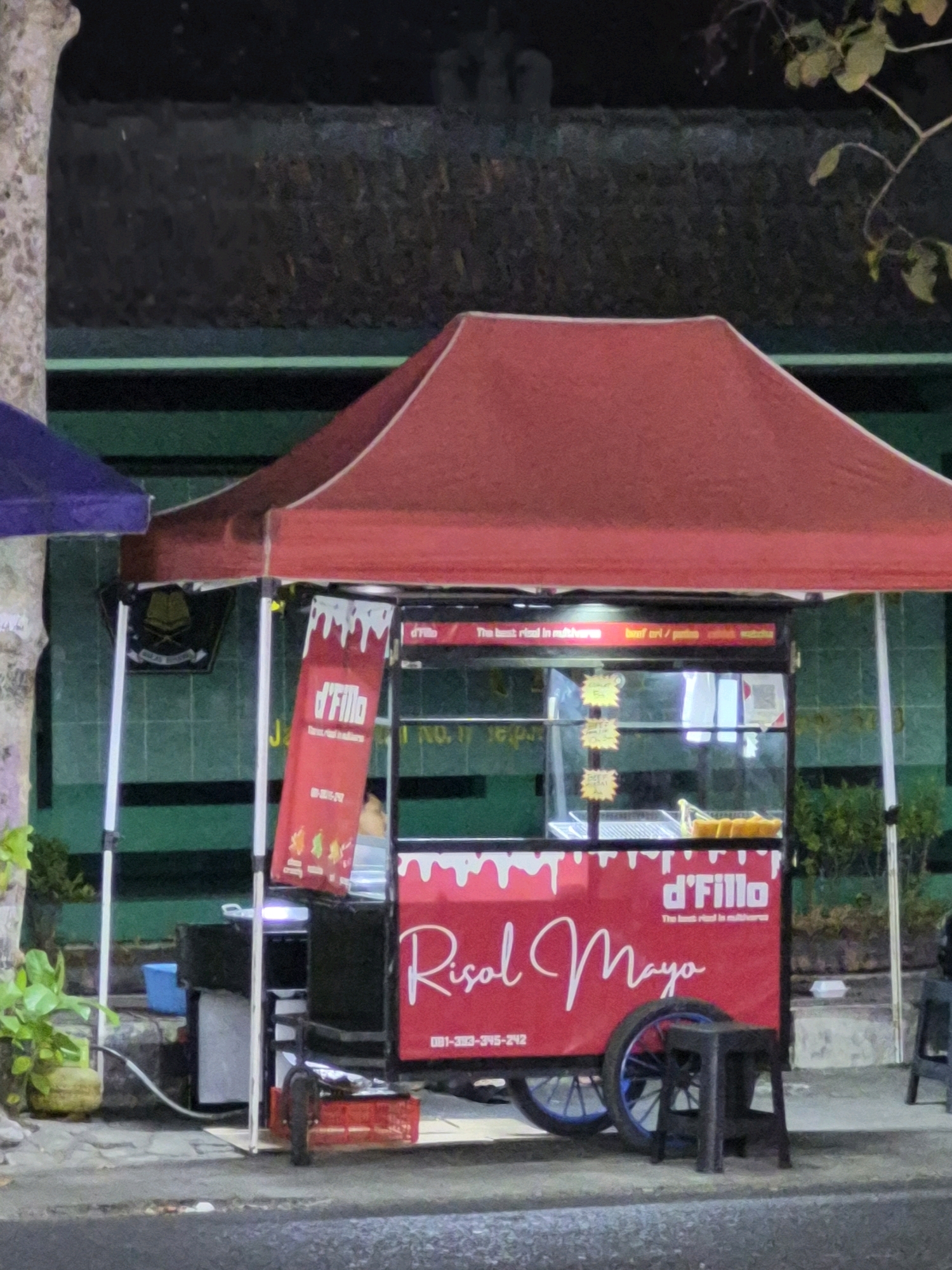 A food cart named "d'Fillo Risol Mayo" is parked on the side of a street at night in Purworejo, Indonesia. The cart has a red canopy and a red front panel displaying the name and a phone number. The cart is equipped with a display case that shows some food items. A black stool is placed in front of the cart. The background shows a green wall with some signage. A tree trunk is visible on the left side of the image. The street in front of the cart appears wet, suggesting recent rain.