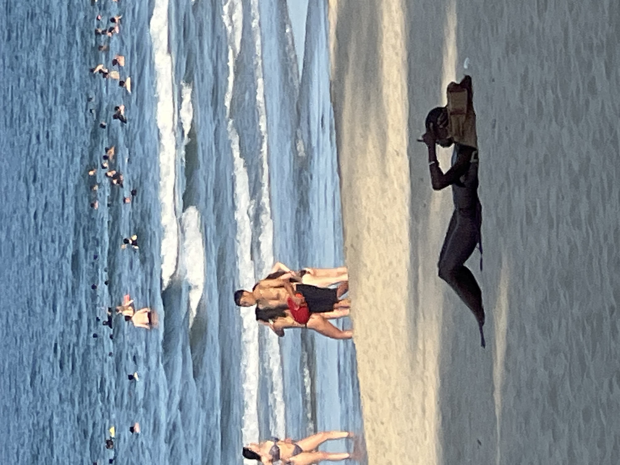 This media file depicts a beach scene in Đà Nẵng, Vietnam. The image is oriented vertically, showing a section of the ocean with waves and a sandy shore. Numerous people are visible in the water, some appearing to swim or wade, while others are closer to the shore.
On the sandy area, a few individuals are present. One person, silhouetted against the brighter sand, is captured in mid-action, holding an object to their ear and face, possibly a phone or camera. Another group of people is shown near the water's edge. One individual is wearing a red object, possibly a lifebuoy or a piece of clothing. Another person is lying on the sand in a bikini. The overall atmosphere suggests a warm, sunny day, typical for a beach outing. The presence of many people in the water and on the shore indicates a popular time for beach activities.