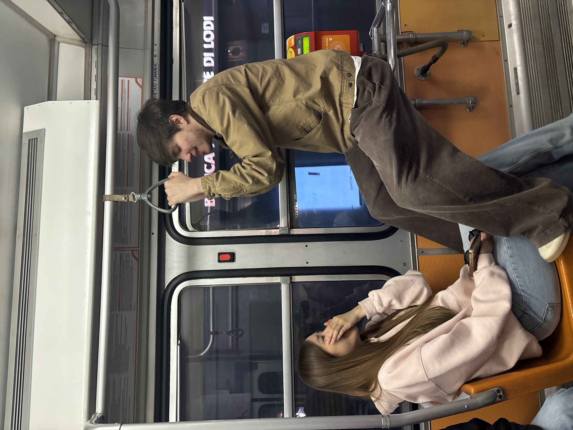 Two young people are inside a train in Milan, Italy. The train has orange plastic seats and grey metal walls. A young man in a tan jacket and brown corduroy pants is leaning forward, holding onto a handle. He appears to be speaking to a young woman seated below, wearing a light pink hooded sweatshirt and jeans. The woman has her hand near her face, possibly covering a smile. Signage on the train includes the words "CA DI LODI." There's a ticket vending machine visible in the background.