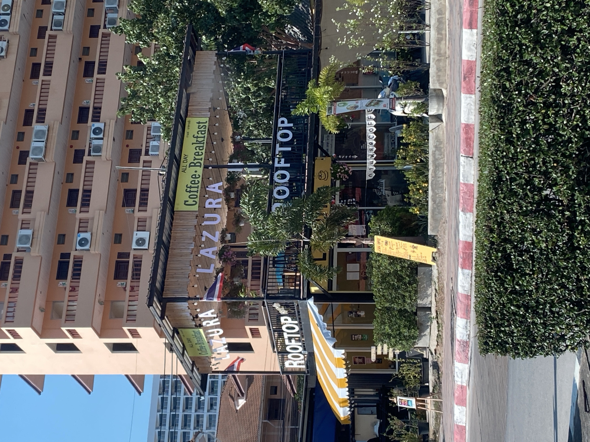 The image depicts the "AZURA LAZURA ROOFTOP" café in Pattaya City, Thailand. The café has both "Coffee.Breakfast" signs and one with "Coffee Breakfast." The café is positioned next to a street bordered by a red and white striped curb and a dense hedge. The café is nestled among buildings, including a tall apartment building with numerous air conditioning units on its façade. Yellow and white striped awnings are visible. Trees and other greenery are around the café, creating a partially shaded environment. The sky is clear and blue in the image's corner. Small flags are also apparent near the café signs. There are also more signs for "กาแฟสดCOFFE" and one in English for "COFFEE."