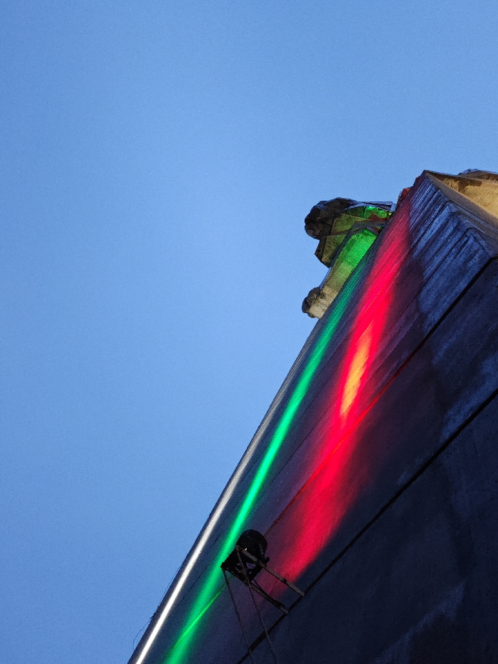 An upward-angled photograph captures a section of the Monument to 1300 Years of Bulgaria (Founders of the Bulgarian State Monument) in Shumen, Bulgaria, against a clear blue sky. The prominent subject is a large, grey concrete facade of the monumental structure, extending diagonally from the lower-left to the upper-right. Two distinct, diagonal beams of artificial light, one green and one red, illuminate the concrete surface. A black spotlight, mounted on a tripod-like stand, is positioned on the facade in the lower-middle section, casting these colored lights upwards. At the very top-right, abstract, angular stone or concrete carvings are visible, including what appears to be a stylized head-like feature. The sky occupies the upper-left quadrant of the image. No individuals are present.