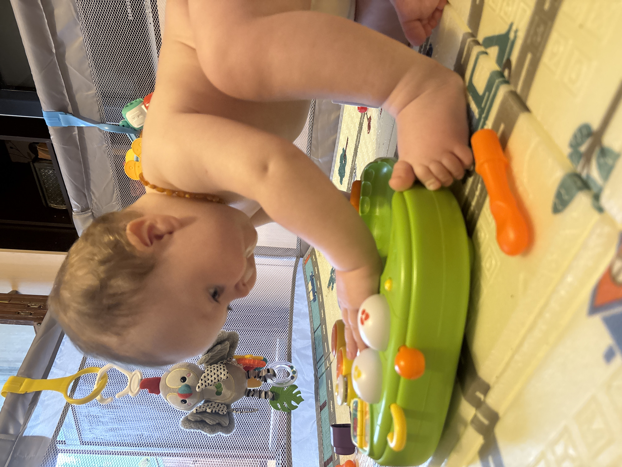 A baby in a diaper is playing with a green and orange toy in a playpen. The playpen is placed on a patterned play mat. There is a mobile hanging inside the playpen. The photo appears to be taken indoors. The location is stated as being in Марковский, Russia, however, no details in the image indicate this as fact.