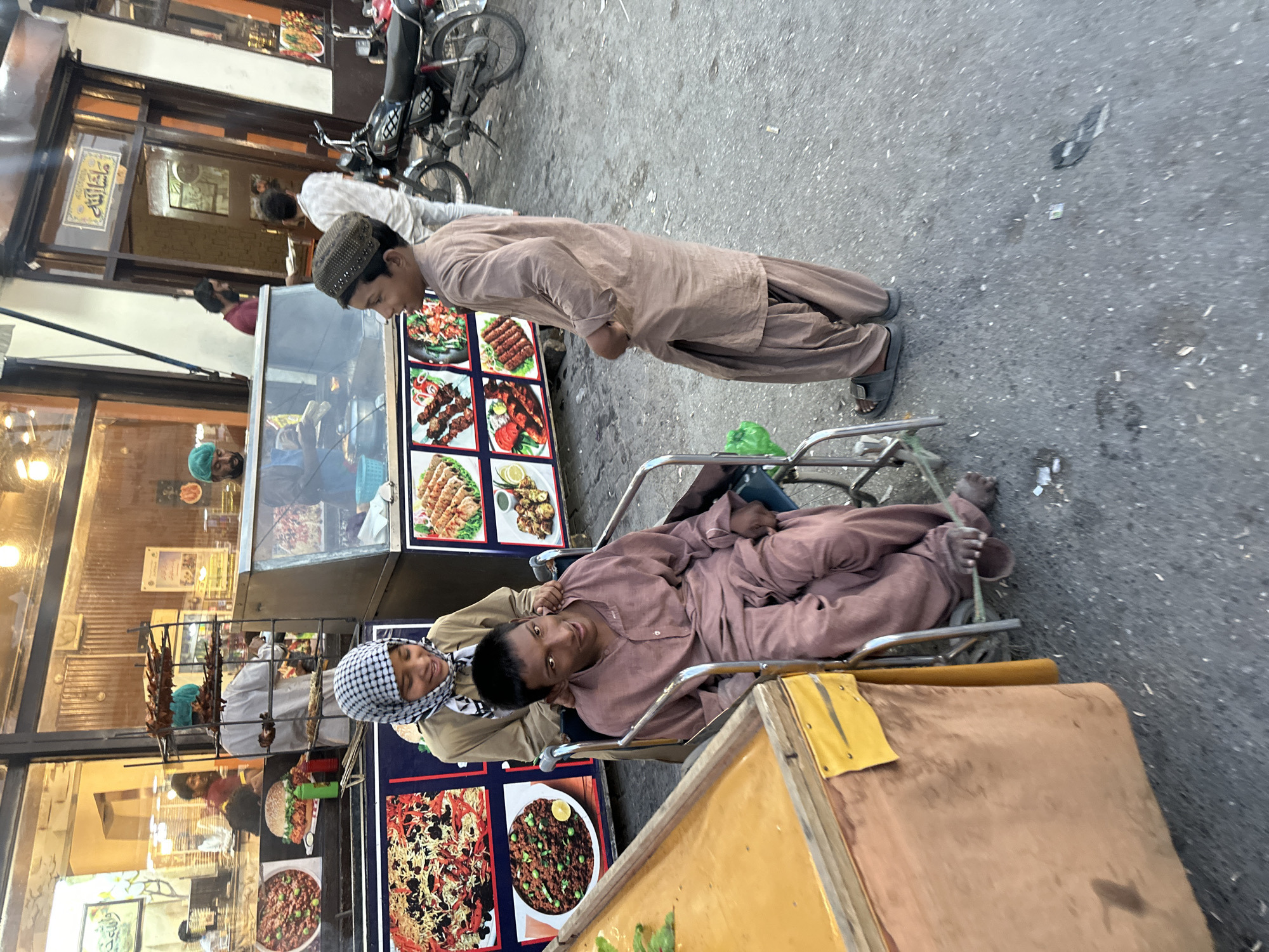 The image captures a street scene in Quetta City Tehsil, Pakistan. A man in a brown salwar kameez is lying in a wheelchair, attended to by another man wearing a checkered headscarf. A third man in a similar outfit stands nearby, observing. The background features a restaurant or food stall with displays of food items. A motorcycle is parked on the street.