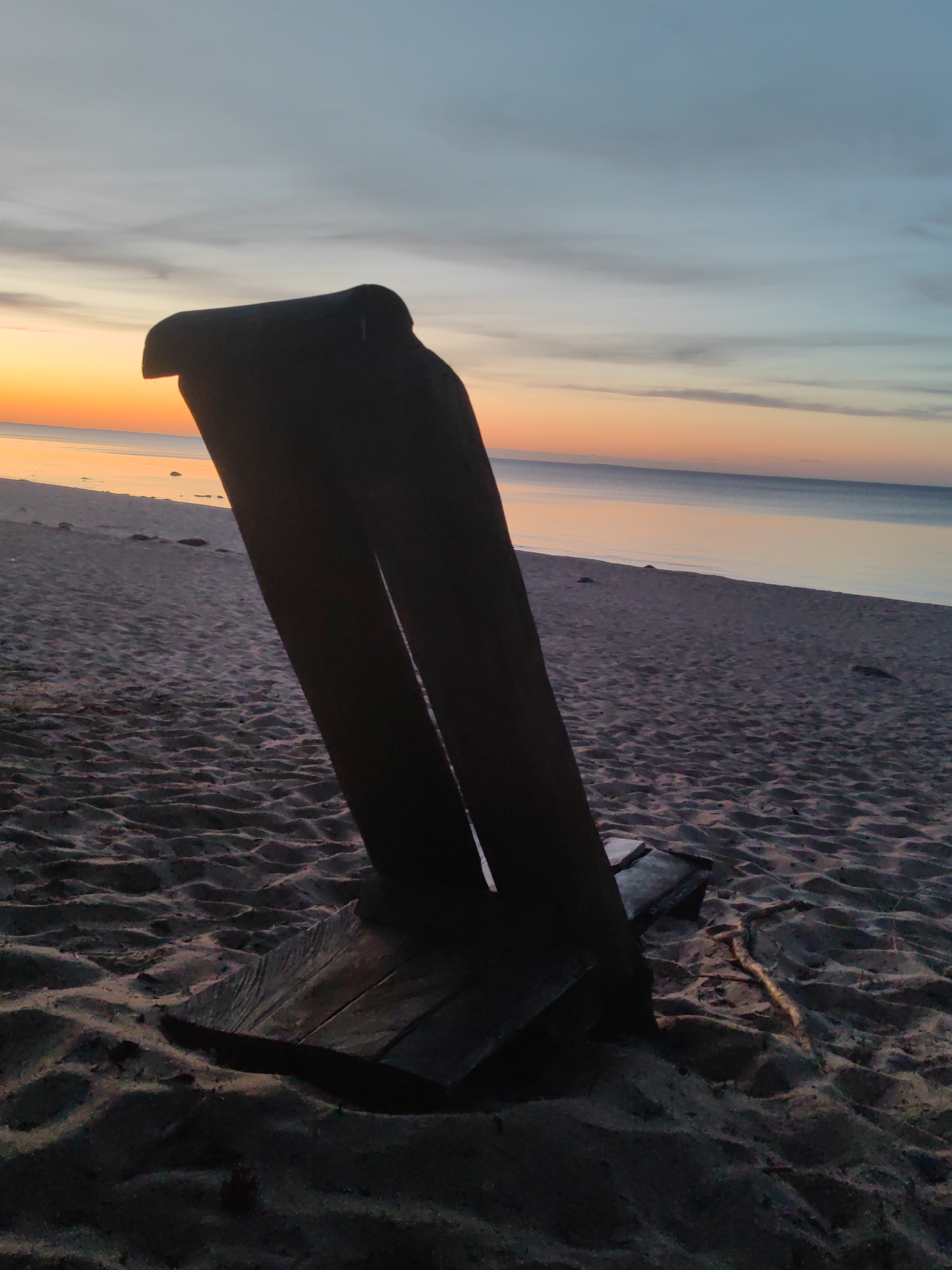 This is a nighttime photograph taken on the beach in Motornoe, Russia. A dark, weathered wooden structure, possibly a sculpture or piece of driftwood, is silhouetted against the twilight sky. The structure is positioned in the foreground, taking up a significant portion of the frame. The beach sand is visible in the lower half of the image, with ripples and indentations indicating its texture. The ocean stretches out in the background, meeting the sky at the horizon. The sky displays a gradient of soft colors, ranging from orange and yellow near the horizon to pale blue and gray higher up, with wispy clouds. There are no people or animals visible in the scene. The overall mood is serene and slightly melancholic due to the fading light and the solitary object on the shore.