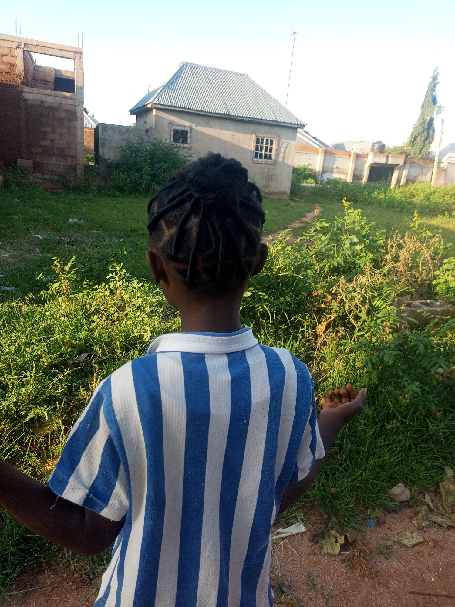 A person, viewed from behind, stands in an outdoor, semi-rural environment with buildings and vegetation. The person has braided dark hair and is wearing a blue and white striped t-shirt. Their right hand is open, palm up, and appears to be holding a small object, possibly a nut or seed. In the background, there are buildings under construction, including one with exposed brickwork and another with a corrugated metal roof. Green grass and various types of vegetation cover the ground, with some debris visible. The sky is clear and bright, suggesting it is daytime. The location is identified as Tempure, Nigeria.