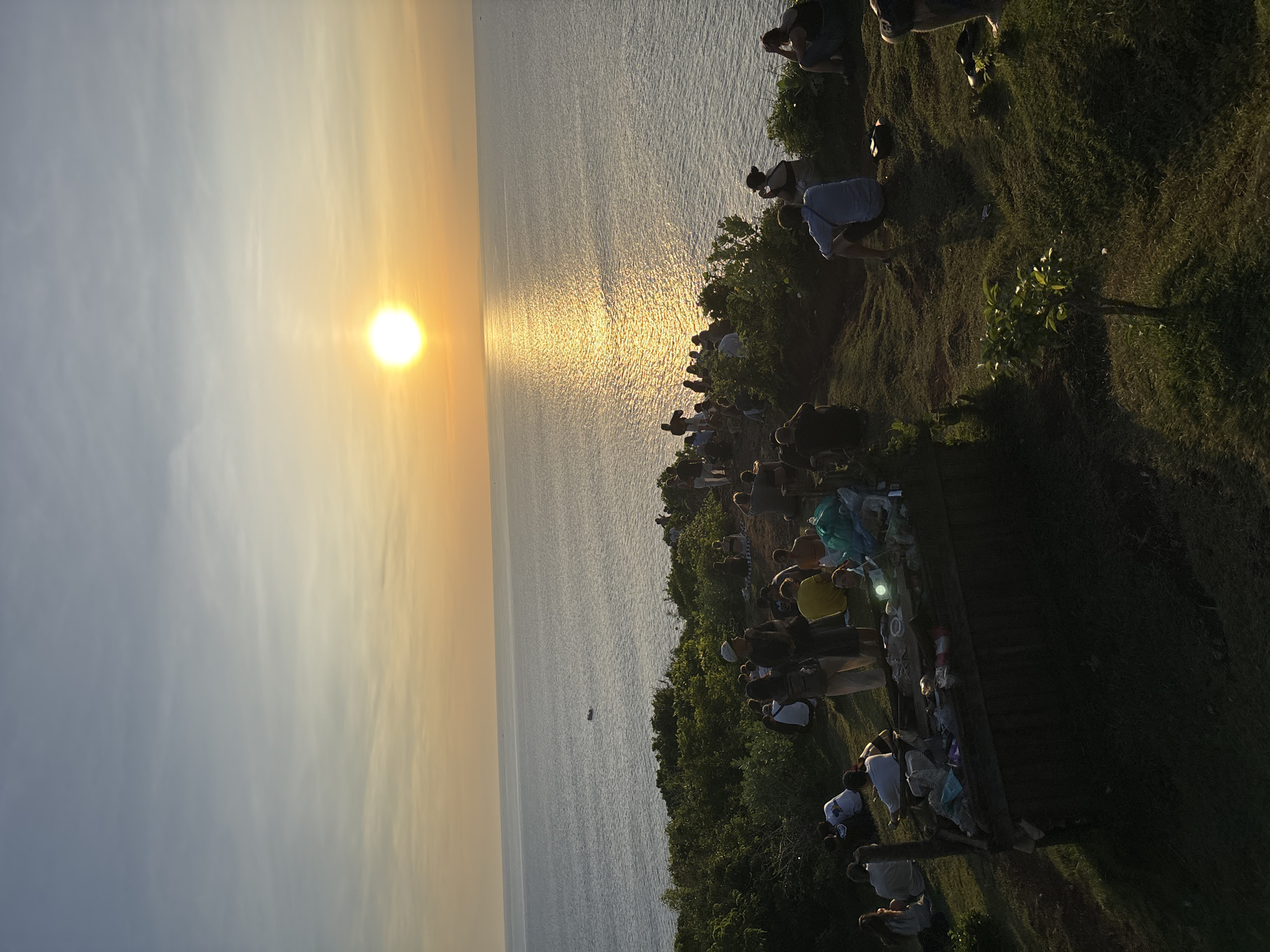 The image shows a sunset view over the ocean, with a crowd of people gathered on a grassy cliff overlooking the water. The sun is low on the horizon, creating a golden reflection on the sea. The location is likely in Kuta Selatan, Indonesia, based on the setting and likely tourism. The people are mostly standing or sitting, looking out at the sunset. There is some vegetation on the cliff, including grass, bushes, and a small tree. A small boat is also visible on the ocean.