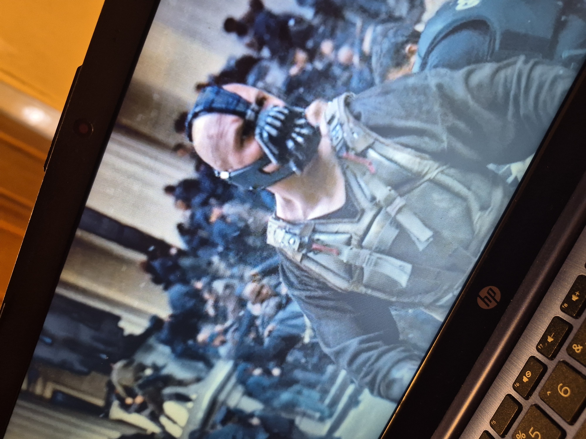 A man wearing a Bane mask and tactical gear is shown on a laptop screen. The laptop has an HP logo and a keyboard with yellow lettering. The background on the screen shows a crowd of people. The image does not provide any information about the location in the city of Sandwell, United Kingdom.