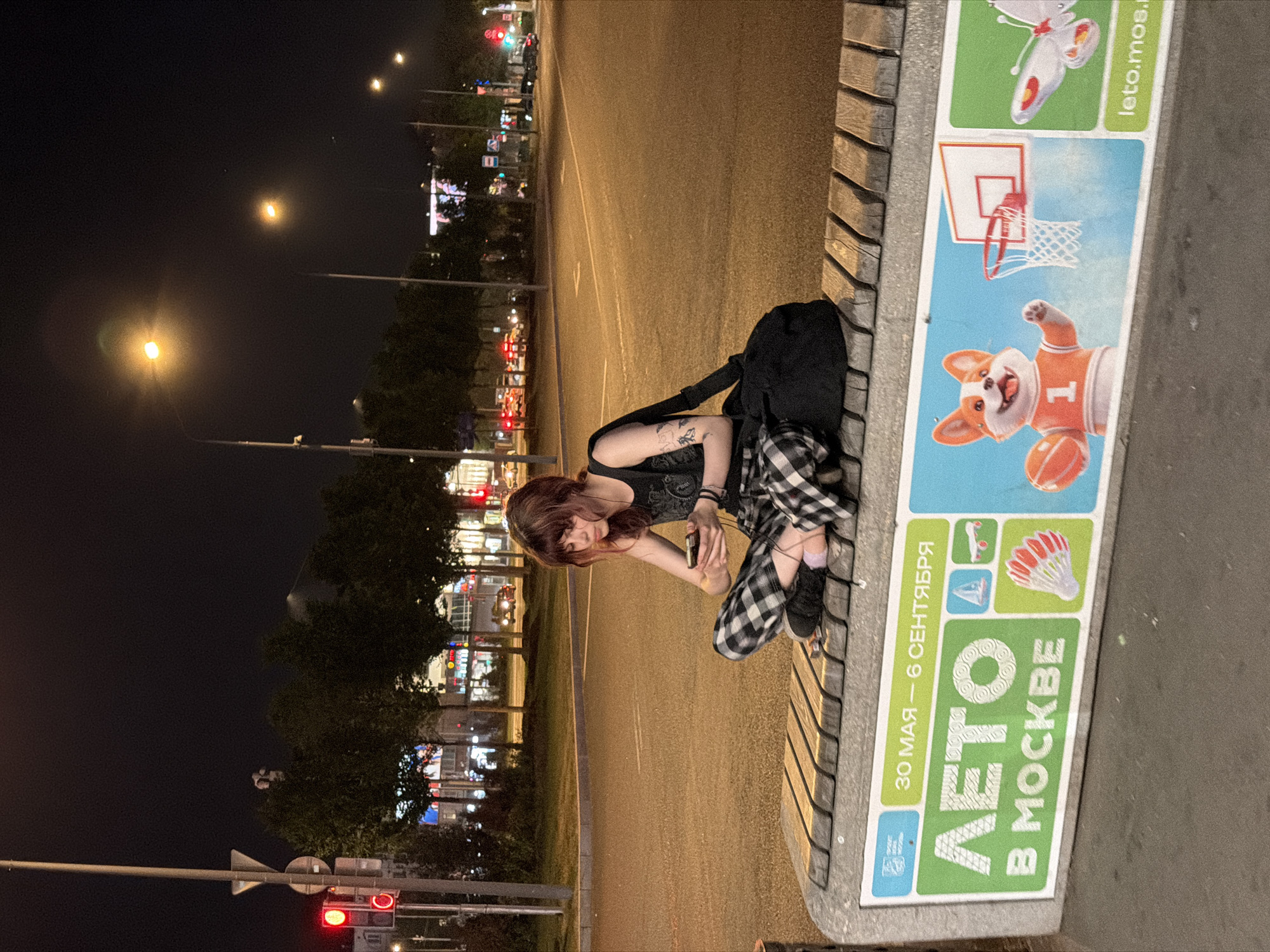 An overhead-level shot captures a young individual seated cross-legged on a public bench at night in Moscow, Russia. The person, appearing to be female with reddish-brown hair and bangs, wears a dark sleeveless top, black and white plaid pants, and dark sneakers with one light purple sock visible. Visible tattoos are present on her left upper arm. She is intently focused on a dark-colored smartphone held in both hands, with earbuds connected and inserted into her ears. A black bag rests beside her on the bench.
The bench consists of concrete supports and wooden slats for seating. Its front panel displays a colorful advertisement in Russian, reading "30 МАЯ – 6 СЕНТЯБРЯ" (May 30 – September 6) and "ЛЕТО В МОСКВЕ" (Summer in Moscow), along with the partially visible website "leto.mos.ru". The advertisement features a logo of the Moscow government and illustrations of summer activities, including a skateboard, a badminton shuttlecock, a cartoon corgi dog in a basketball jersey holding a basketball next to a hoop, and a butterfly.
The background depicts a wide, multi-lane asphalt road with visible lane markings. Numerous streetlights with yellow-orange illumination line the thoroughfare, and a red traffic light is visible on the left side, accompanied by a pole displaying a yield sign. In the distance, several vehicles with illuminated headlights and taillights indicate ongoing traffic. Behind the road, trees and various illuminated buildings suggest an urban environment. The sky is dark, characteristic of nighttime.