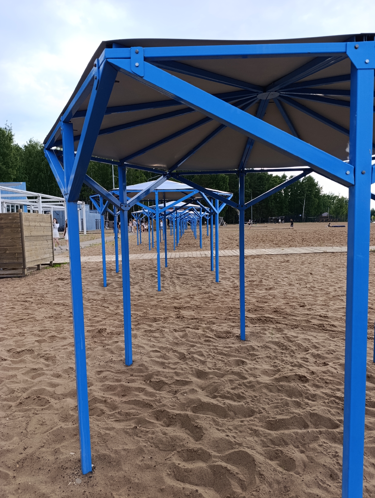 An outdoor shot captures a row of blue, octagonal beach shelters on a sandy beach. The shelters are constructed with blue metal poles supporting beige fabric tops. The perspective is from within one of the shelters, looking down the line of similar structures. The sand is textured with footprints and ripples. In the background, a wooden changing room or structure is visible on the left, with a few people walking near it. Further back, trees form a treeline, and a few people can be seen scattered across the sandy expanse, some engaged in activities. The sky is overcast.