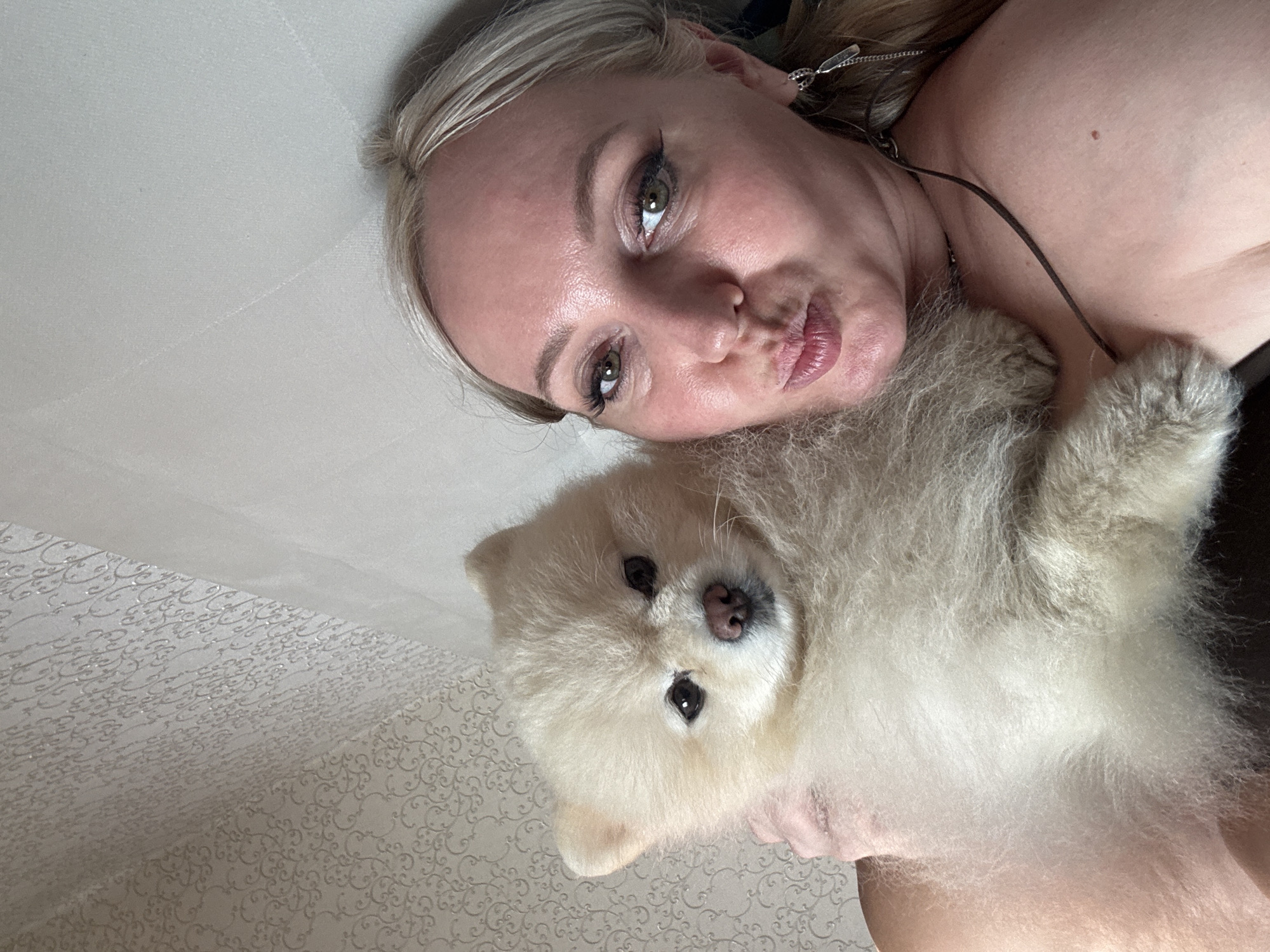 A close-up selfie shows a woman with blonde hair and light-colored skin holding a fluffy Pomeranian dog. The woman is looking at the camera with her lips puckered. She is wearing makeup, including eyeliner and mascara. A silver earring dangles from her right ear. The dog is cream-colored with dark eyes and a pink nose. They are positioned in front of a wall with textured wallpaper featuring a repeating pattern. The lighting suggests an indoor setting, possibly a home in Komsomolsk-on-Amur, Russia.