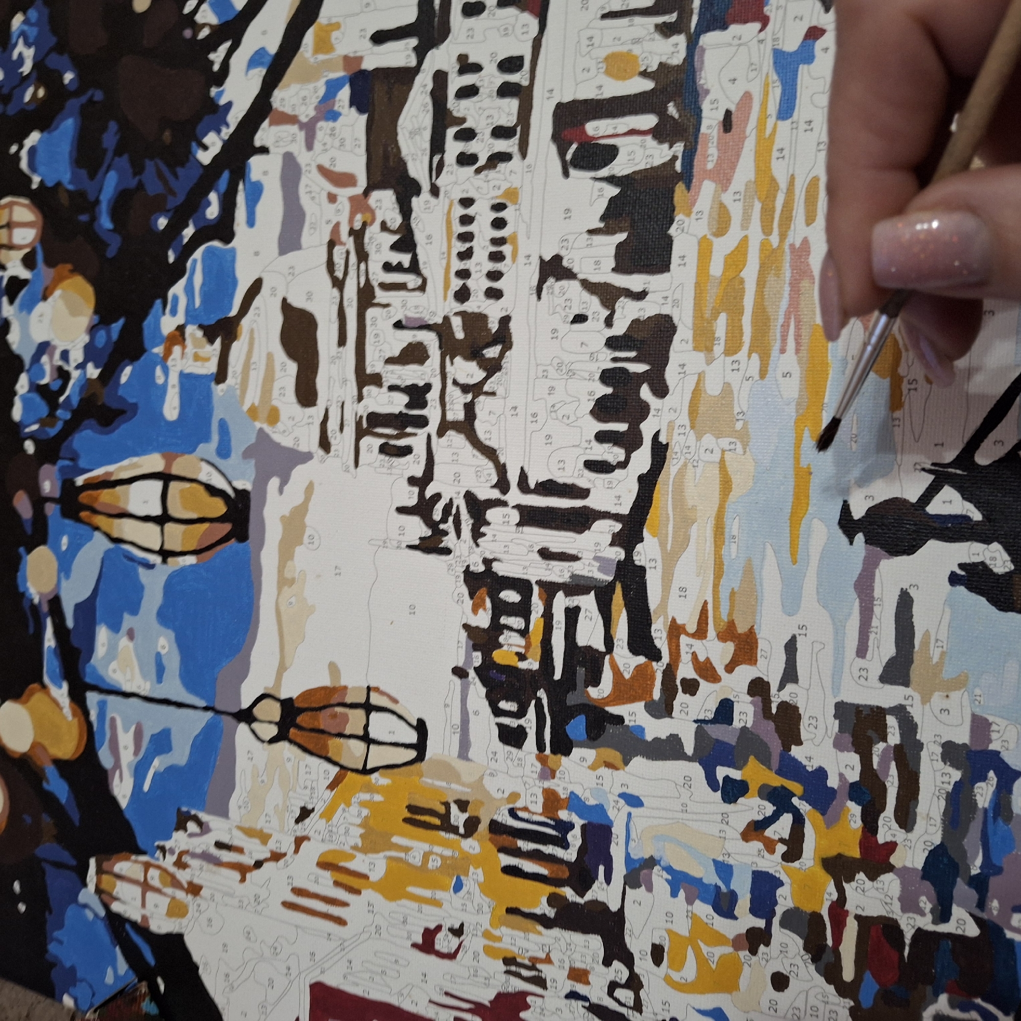 A close-up shot depicts a canvas with a paint-by-numbers design, specifically of a city scene. The canvas is partitioned into various numbered sections, indicating the colors to be applied. A hand holding a paintbrush is visible, poised to paint a section of the canvas. The canvas depicts buildings, street lights, and sky in various shades of yellow, brown, blue, and black. It's apparent that some areas of the canvas are already painted. The style of art appears to be impressionistic, capturing the essence of the city rather than a hyper-realistic rendering. The location, city, or people are not identified.