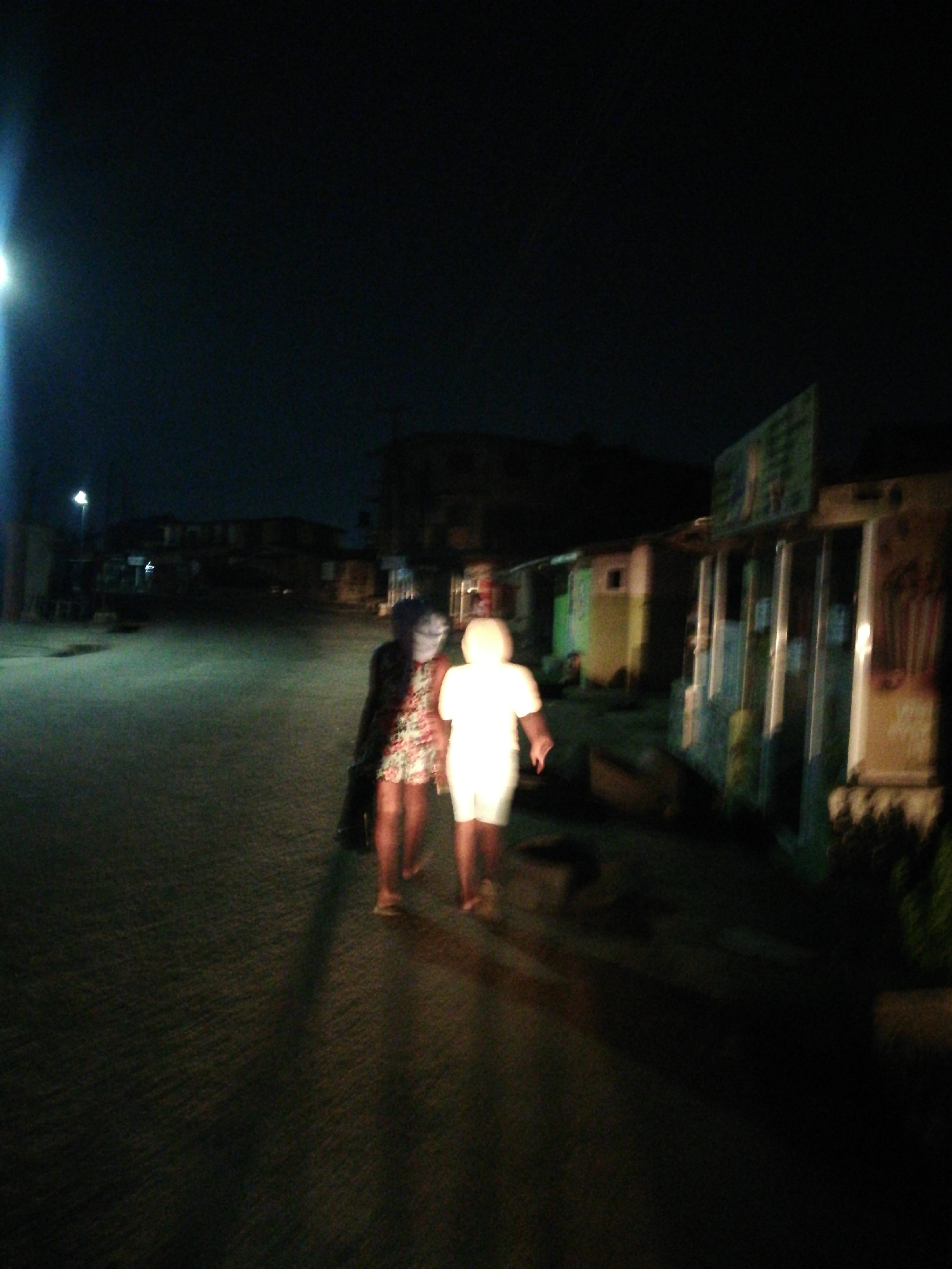Two figures walk down a dimly lit street at night in Lagos, Nigeria. One figure wears a floral dress and dark jacket. The other wears a white outfit. Buildings line the right side of the street. A street light is visible in the distance.