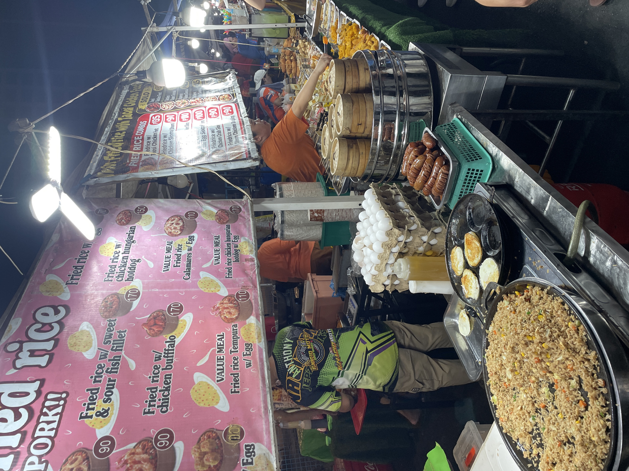 The image depicts a street food stall in Davao City, Philippines, specializing in fried rice dishes. The stall features a vibrant pink banner displaying menu items like "Fried rice w/ sweet & sour fish fillet" and "Fried rice w/ chicken buffalo," along with prices. Another banner advertises "Twin Fried Rice Combos with Taosi Chicken Feet."
In the foreground, a large pan of freshly cooked fried rice is visible. Next to it, a griddle is cooking several small, round fried eggs. A tray of sausages is also displayed in a green basket, alongside a stack of egg cartons. A vendor, wearing an orange shirt, is attending to the food preparation. Another person, wearing a colorful shirt with the name "Anthony" and "Ignacio Moto" printed on it, stands behind the counter, appearing to be a worker. The stall is illuminated by overhead lights, and various food items, including skewers of meat and vegetables, are arranged on display tables behind the vendor. The overall scene captures the bustling atmosphere of a night market or street food establishment.