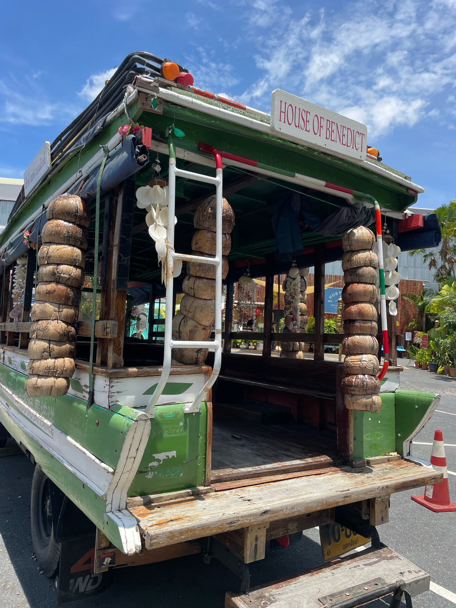 The image shows a side view of a brightly painted, green and white open-air bus with a wooden flatbed, parked on asphalt in an outdoor setting. The bus is decorated with numerous strings of stacked coconuts hanging from its sides, as well as strings of shells. A white sign with red lettering reading "HOUSE OF BENEDICT" is affixed to the top of the bus. The bus has a ladder attached to its side. In the background, there are palm trees, other vegetation, and some buildings, suggesting a tropical or subtropical environment. A red and white traffic cone is visible to the right of the bus. The sky above is blue with scattered clouds. The bus appears to be in a state of disrepair, with peeling paint and weathered wood. The overall impression is that of a unique, possibly tourist-oriented vehicle. The license plate visible on the back of the bus contains Thai script and numbers. While the exact location within Pattaya City is not identifiable from the image alone, the presence of Thai script and tropical vegetation strongly indicates it is in Thailand, likely in or around a tourist area. There are no people visible in the image.