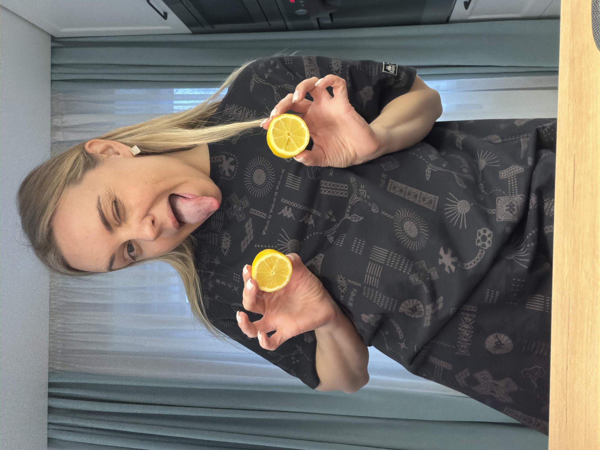 A woman with blonde hair and fair skin is holding two halves of a lemon. She is wearing a black shirt with a pattern of various symbols. She is sticking out her tongue and winking. The background includes a window with light blue curtains, a cabinet or appliance, and a light brown wooden surface. The location is not specified, and there is no evidence to suggest the image was taken in Votkinsk, Russia.