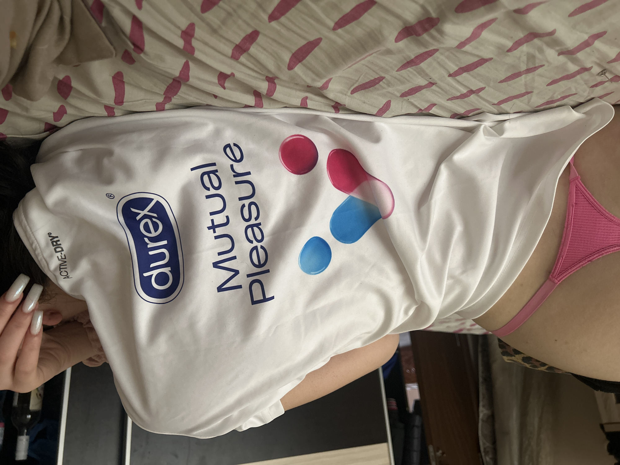 The image shows a person lying on a bed with a pink patterned sheet. The person is wearing a white T-shirt with the Durex logo and the words "Mutual Pleasure" printed on it. A pink thong is visible on the person's lower body. Long, manicured nails are visible on the person's left hand. A dark object, possibly a bottle, is also partially visible in the lower-left corner. The background includes a dark surface, possibly a piece of furniture. The location is not identifiable from the given image.