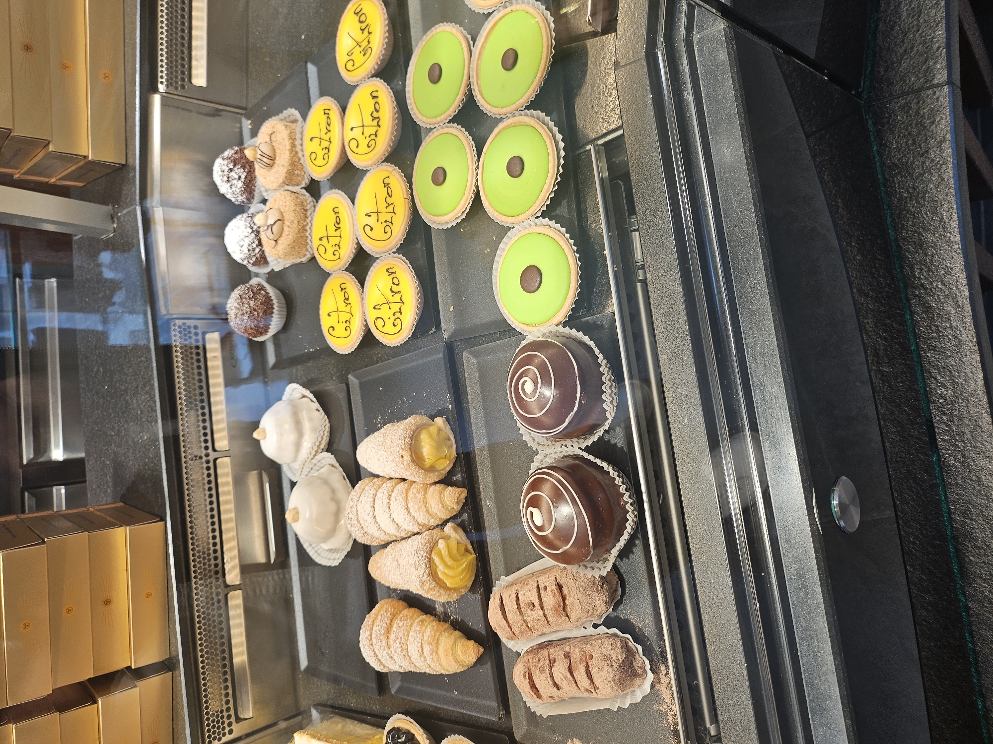 The image displays a selection of pastries in a display case, likely in a bakery or patisserie in St. Moritz, Switzerland. The pastries are arranged on black trays. In the top section, there are round tarts with yellow filling, some with "Guitron" written in black script on top, and others with bright green filling topped with a single chocolate piece. Below these are several cream-filled pastries shaped like cones, dusted with powdered sugar. There are also chocolate-covered pastries with white swirl decorations and dark chocolate pastries dusted with cocoa powder.
To the left of the pastries, stacked golden boxes with embossed designs are visible, suggesting packaged goods for sale. The overall setting appears to be indoors, with the display case reflecting the surroundings. The lighting suggests it could be daytime. There are no people visible in the frame. The context of St. Moritz, Switzerland, implies a potential setting in a resort town, possibly catering to tourists. The focus is solely on the visually appealing arrangement of the baked goods.