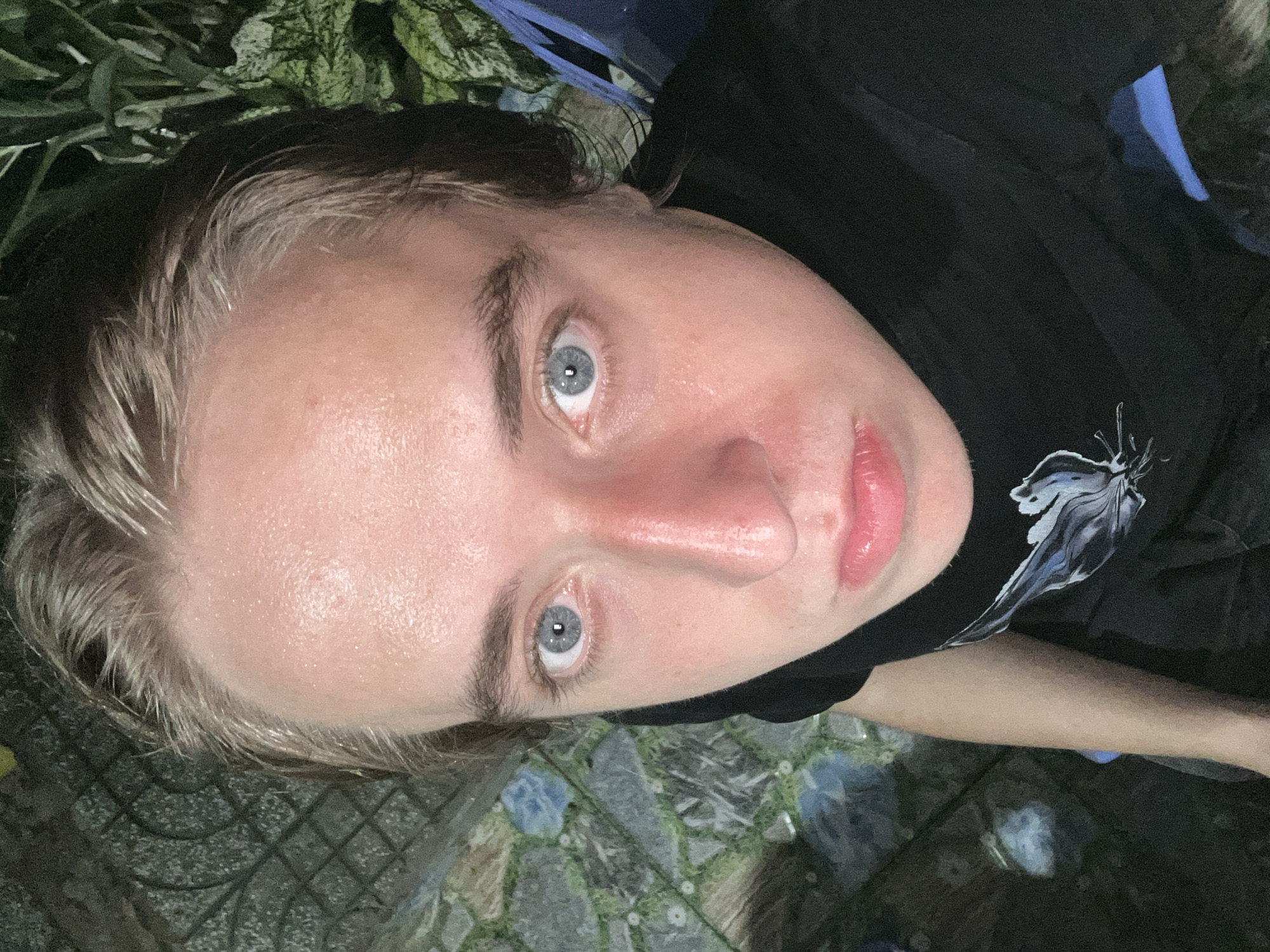 A high-angle, medium shot captures a young person with fair skin and light brown hair, wearing a black t-shirt, as they look directly at the camera. Their eyes are a striking blue. The skin on their forehead and cheeks appears to be glistening with sweat. The background consists of a patterned stone-tiled ground and dark foliage. Hints of a blue chair are visible on the right side of the frame. The lighting suggests an outdoor setting at night. The location is identified as An Hữu, Vietnam.