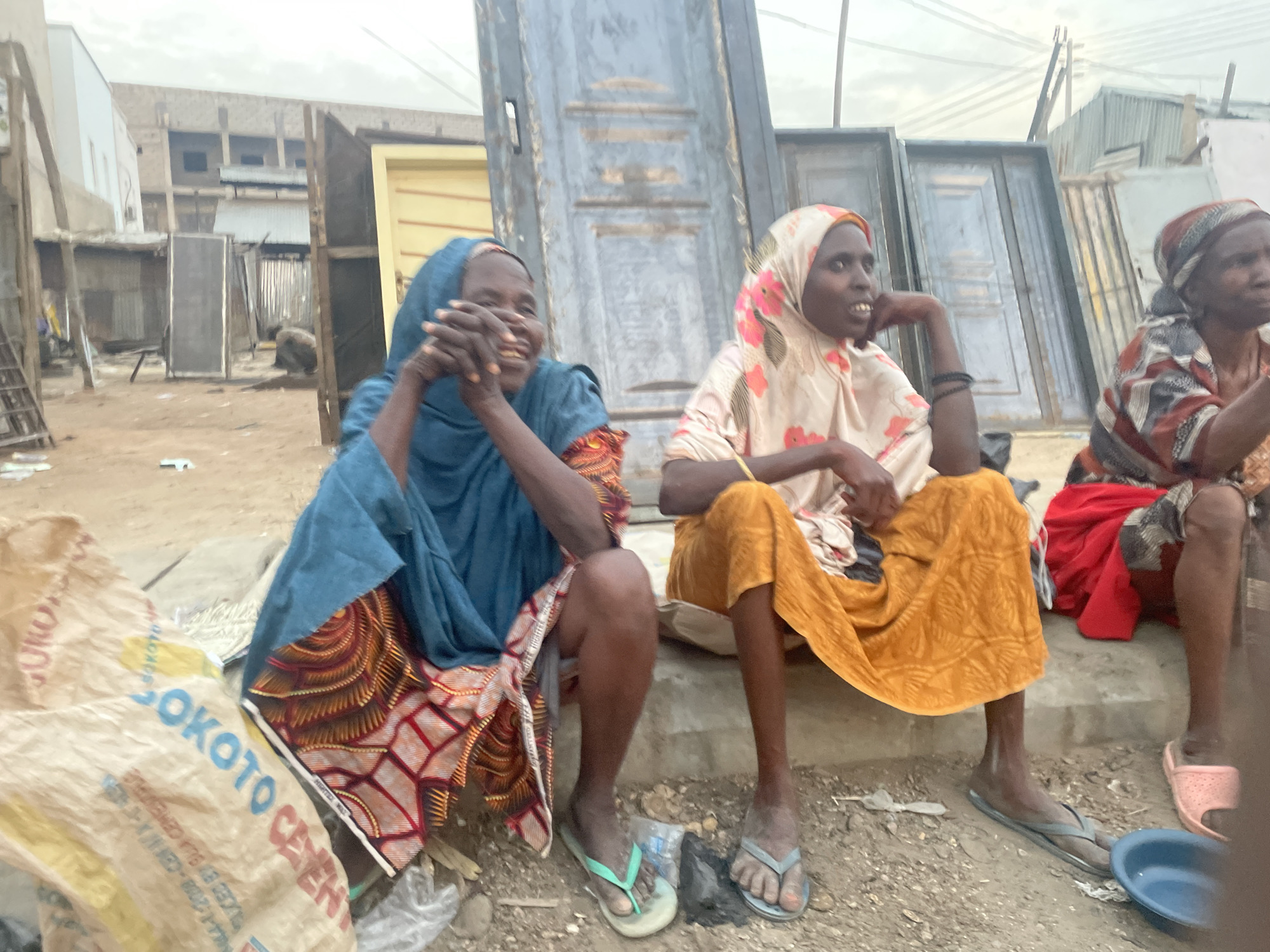 Three women are seated on a low concrete structure in an urban environment, likely a public space or market in Unknown, Unknown. In the background are several doors leaning against each other. The woman on the left is wearing a blue head covering and a colorful patterned dress, smiling as she holds her hands together. The woman in the middle wears a floral headscarf and a yellow dress, with her arm resting on her knee. The woman on the right wears a patterned top and red fabric, appearing older with a serious expression. In the foreground is a large, partially obscured bag labeled "SOKOTO CEMENT." The ground is dirt and gravel, with various objects scattered around. Buildings and structures are visible in the background.