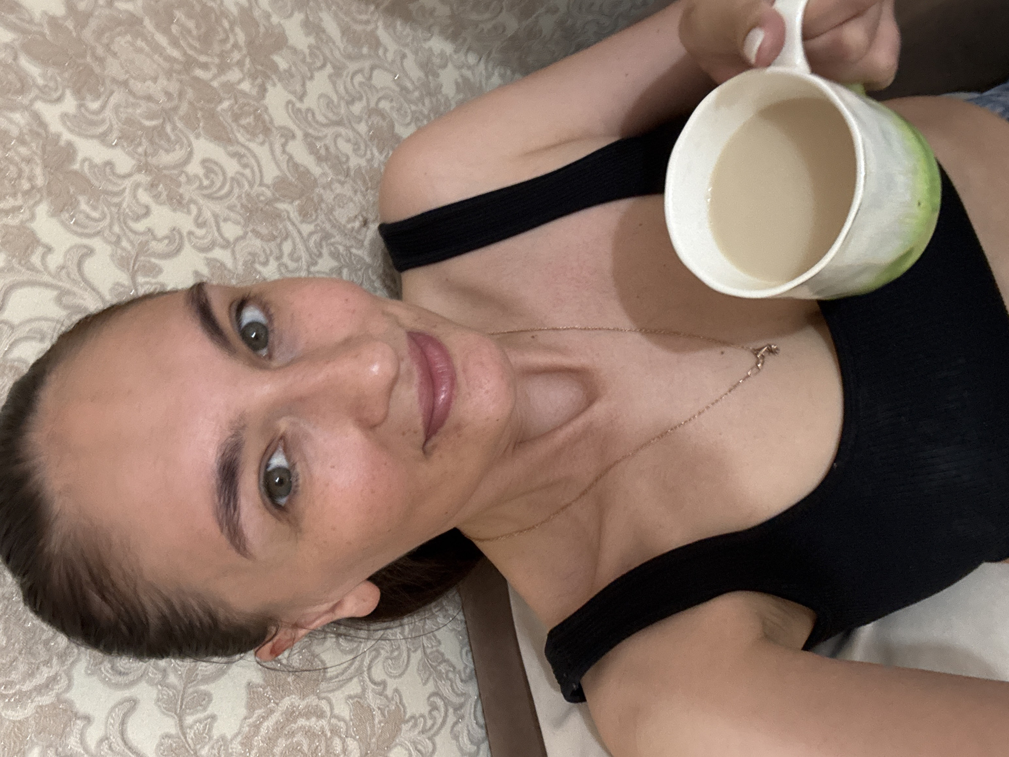 A woman is lying on a bed with a patterned beige duvet, holding a mug filled with a light-colored beverage. She is wearing a black top and a delicate gold necklace. Her face is angled toward the camera. The location in the city of Sochi, Russia, cannot be determined from the image.